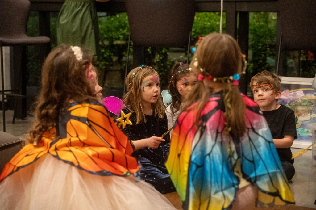 A group of children in costumes and face paint sit in a circle indoors, appearing to participate in a group activity or conversation.