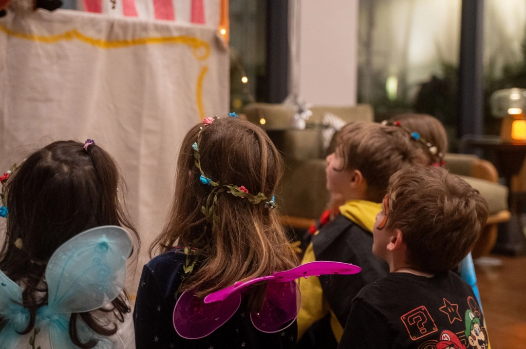 Several children dressed in costumes and fairy wings sit closely together, facing a stage with a curtain, watching a performance indoors.