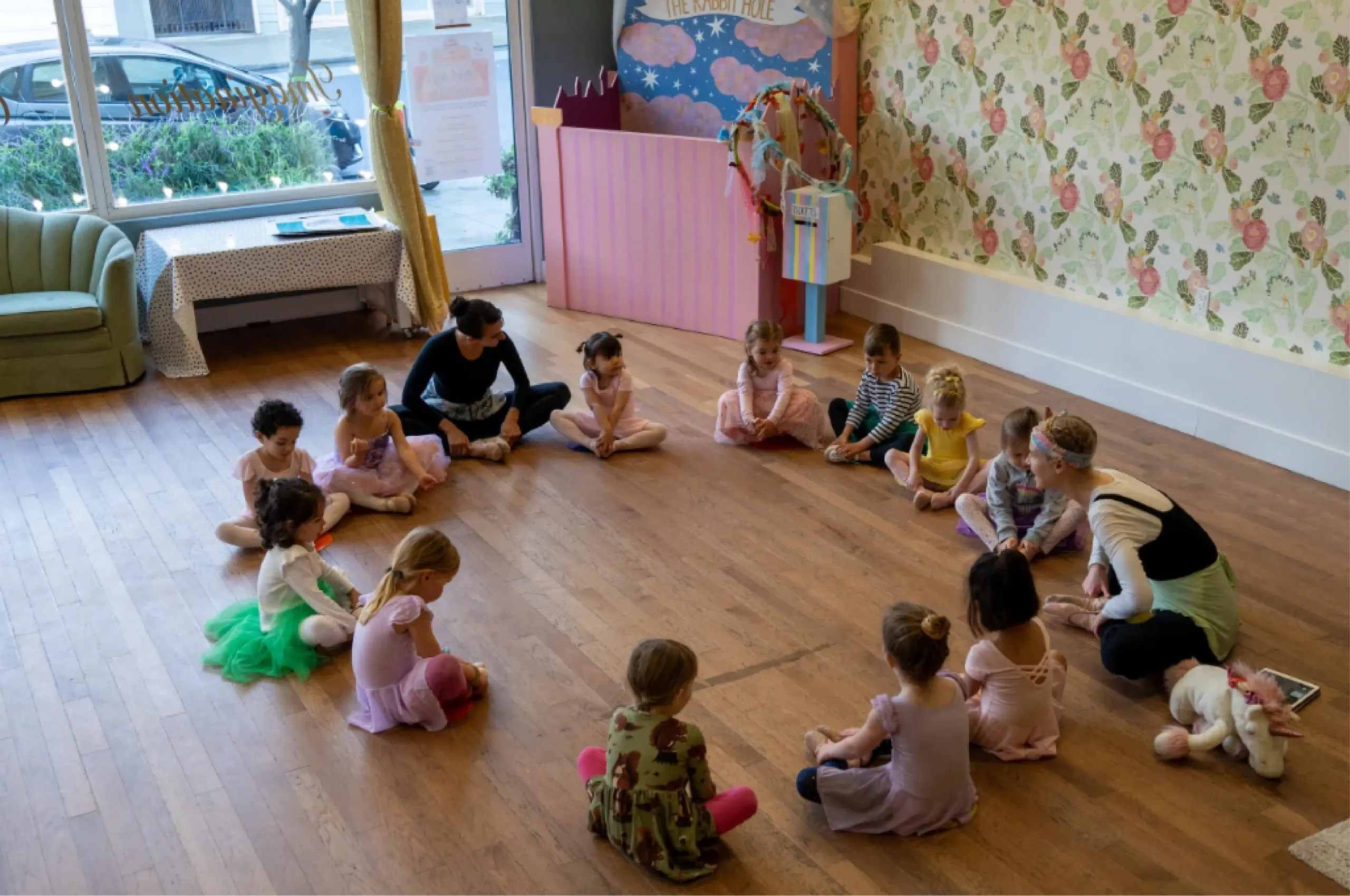 A group of young children in ballet outfits sit in a circle on the floor of a decorated dance studio with two adults.