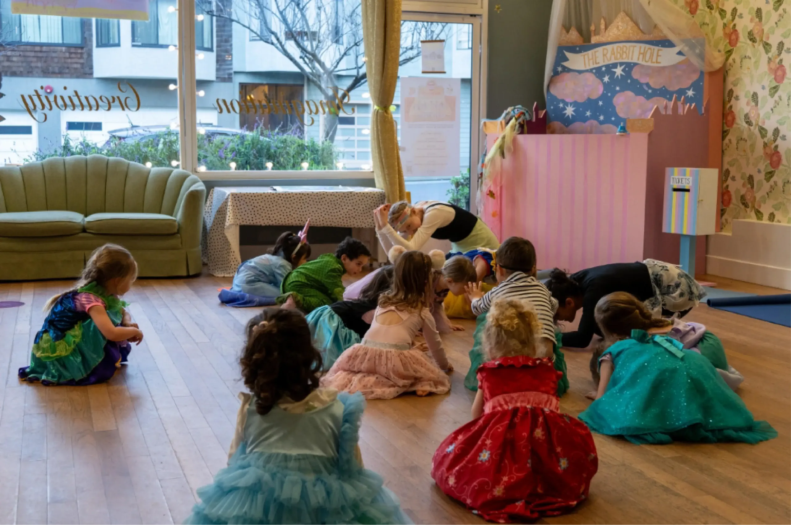Children in colorful costumes sit and lie on the floor in a decorated room, participating in an organized group activity led by an adult.