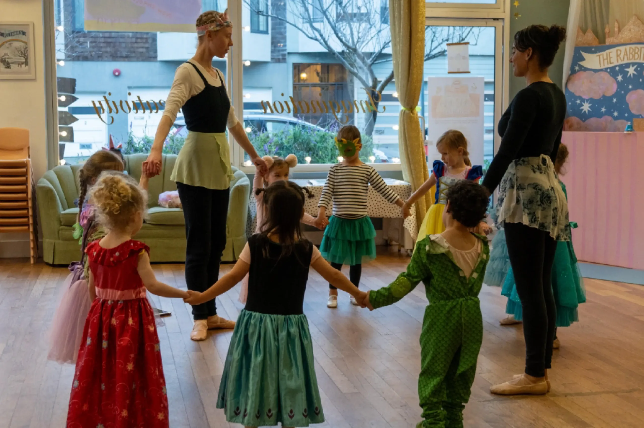 Children dressed in costumes hold hands in a circle with two adults in a dance studio, participating in a group activity.