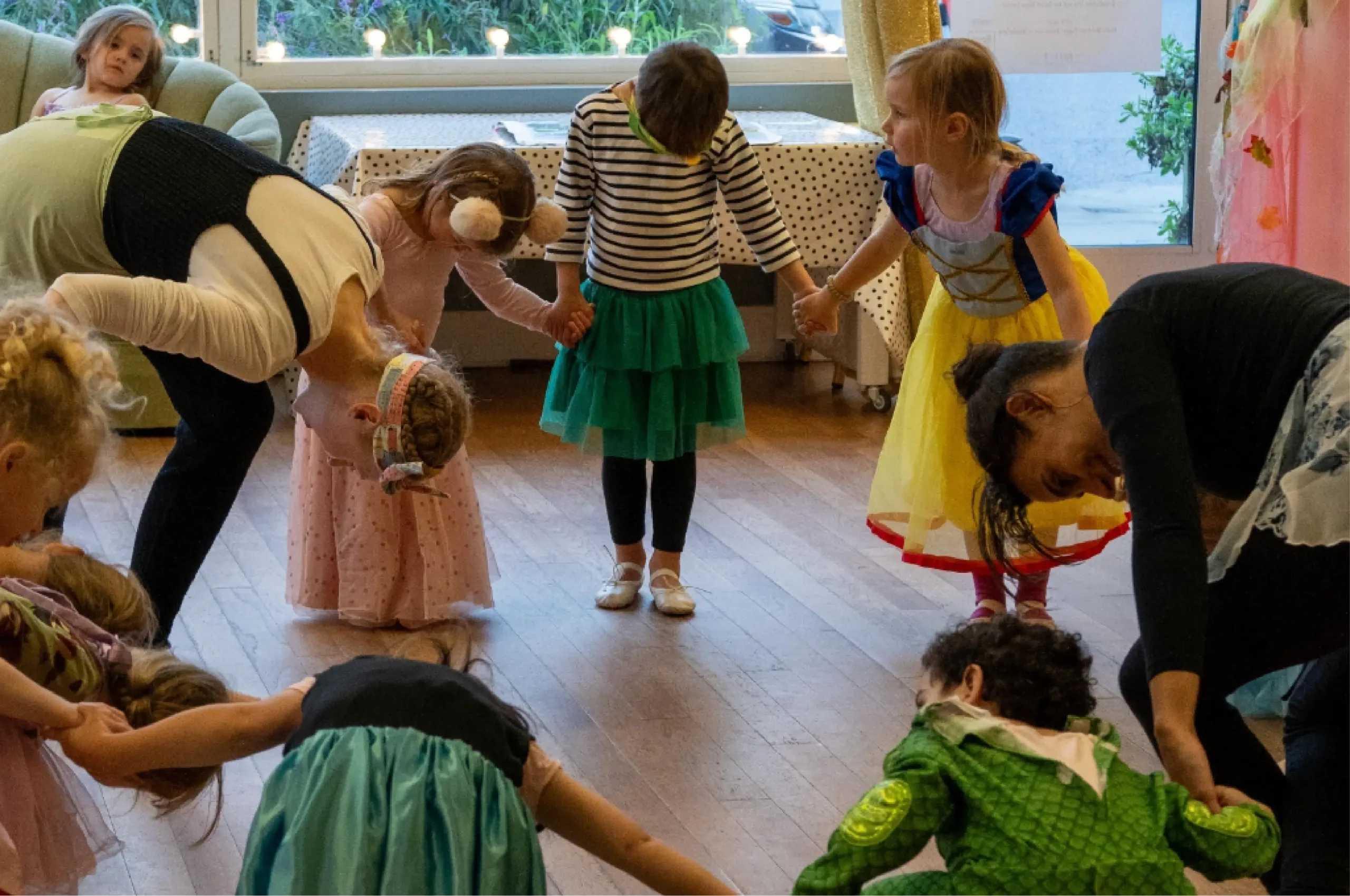 Children and adults in costumes stand in a circle holding hands and bending forward in a room with wooden floors and large windows.