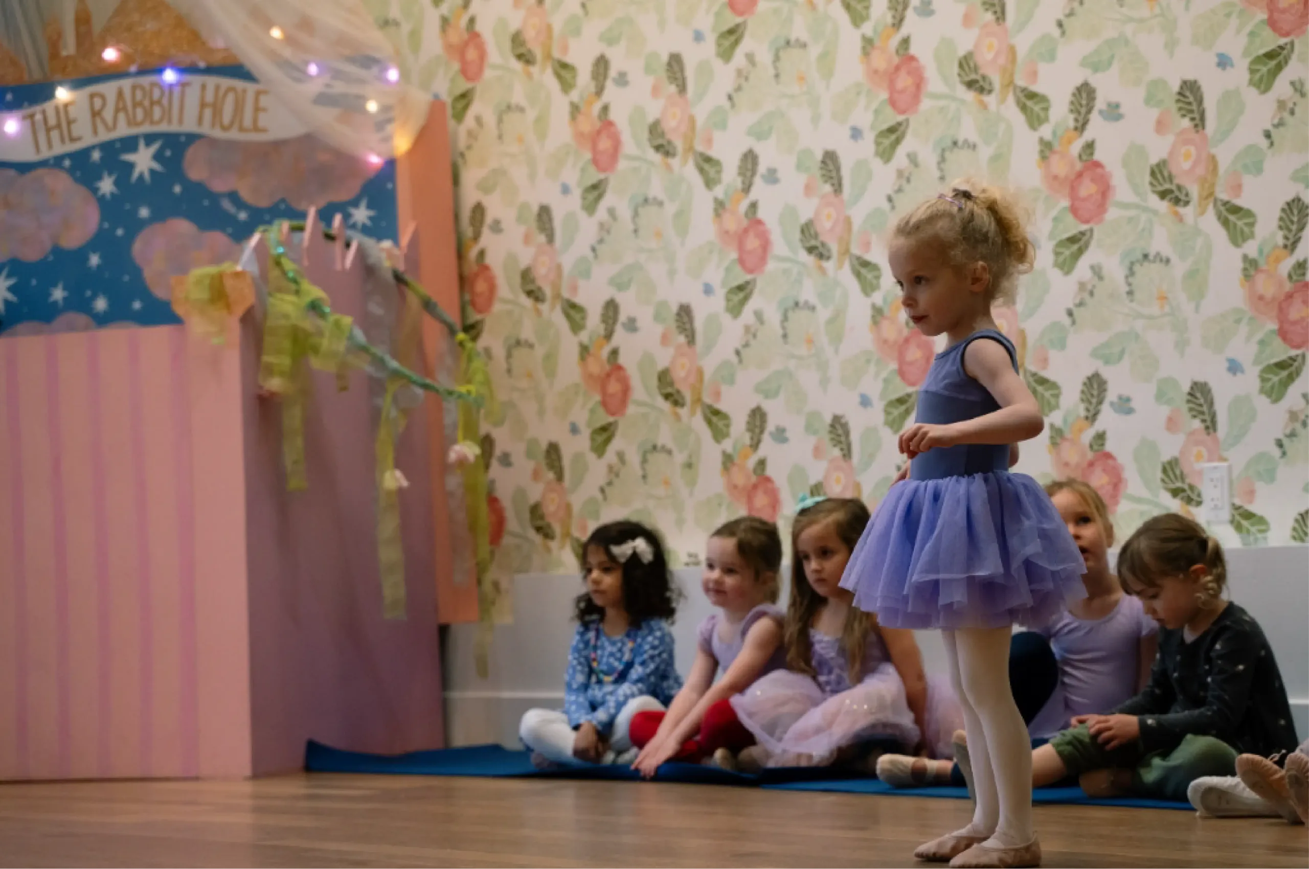 A young girl in a purple ballet outfit stands poised on one foot while other children sit on the floor in a decorated room with floral wallpaper.