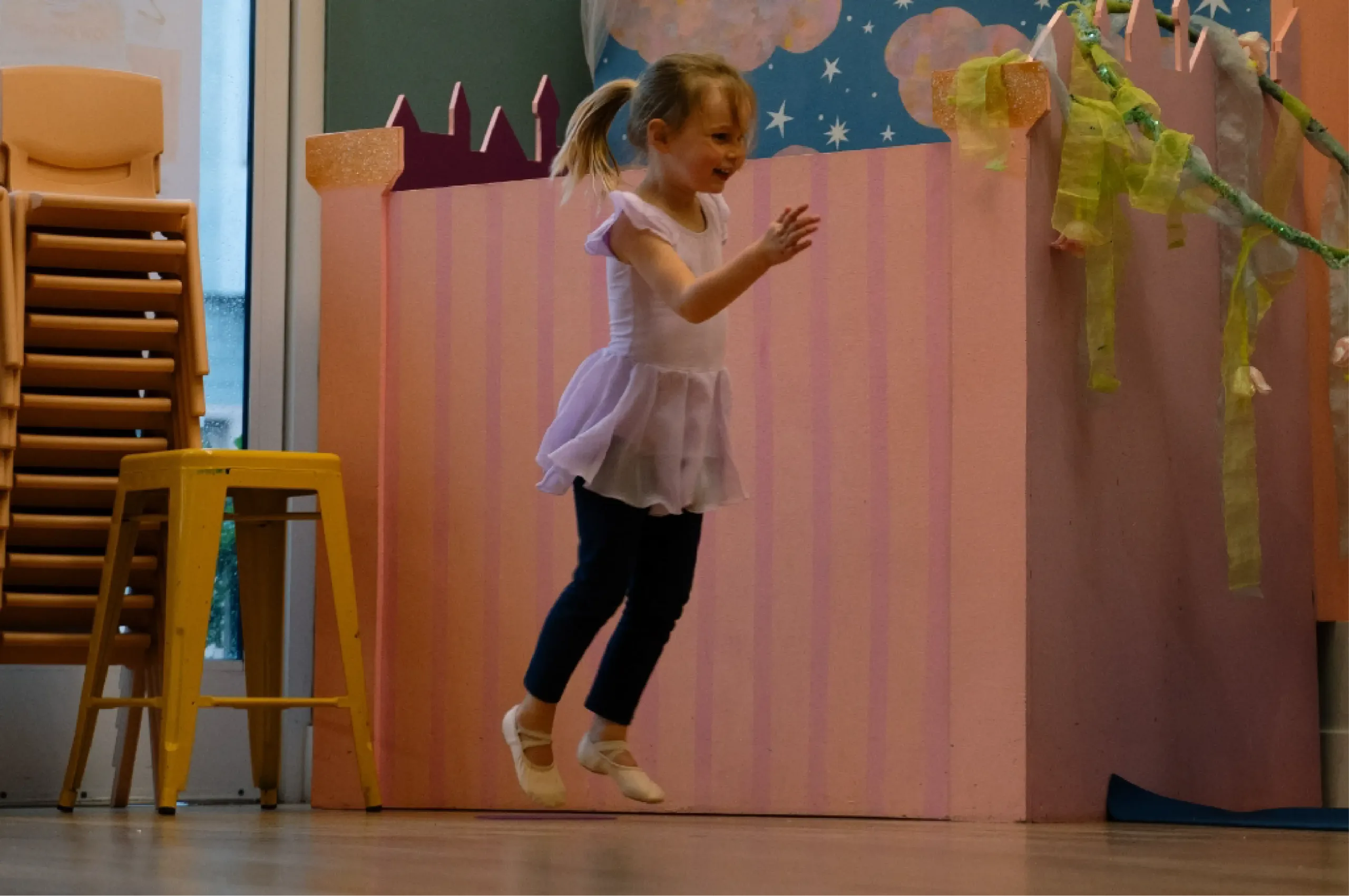 Young child in ballet attire jumps in a dance studio with pastel decorations, including a yellow stool, stacked chairs, and a pink striped backdrop.