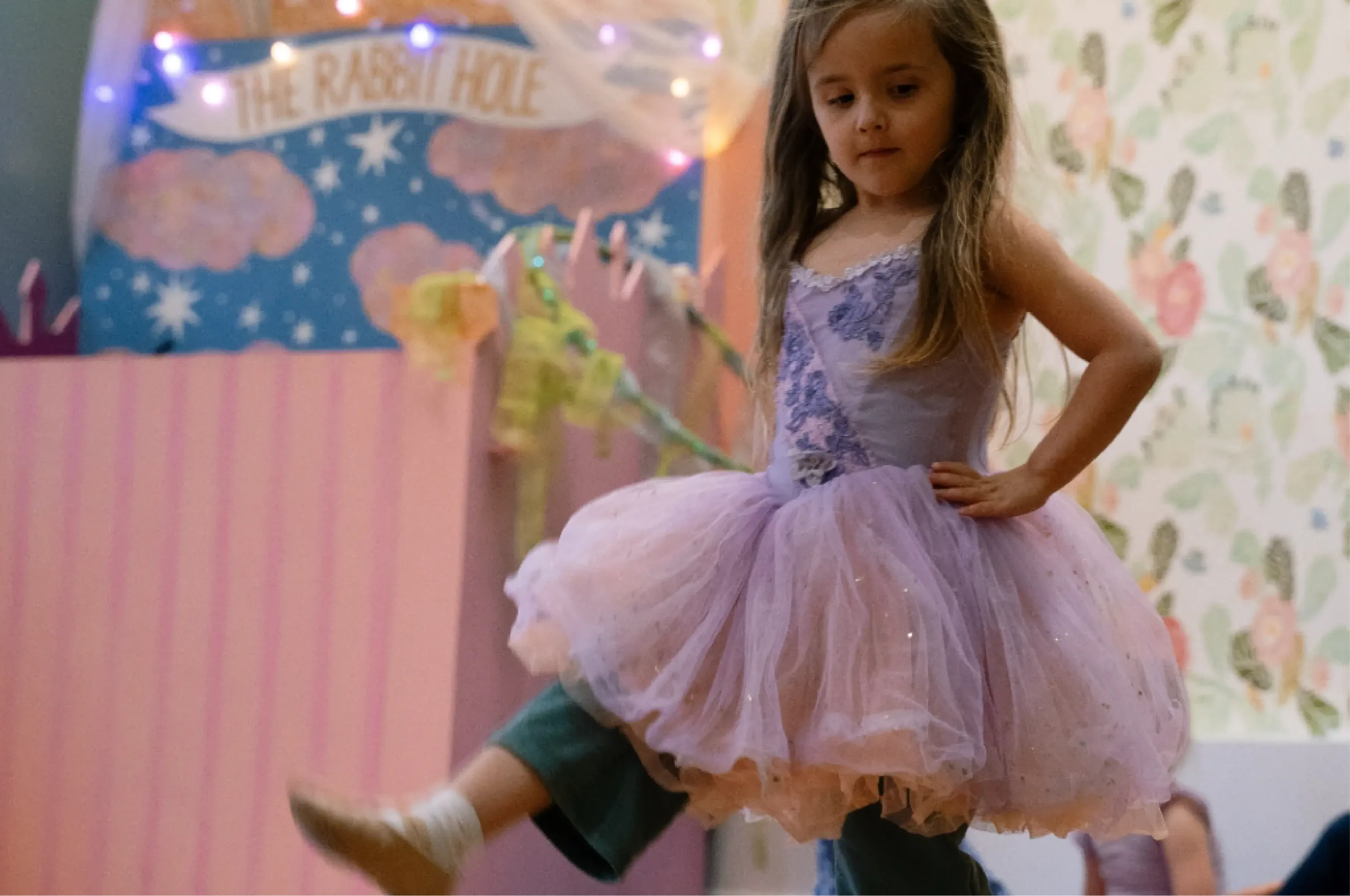 A young girl wearing a lavender tutu and green pants dances indoors, with one leg raised and her hand on her hip. The background has pastel decorations and a "The Rabbit Hole" sign.