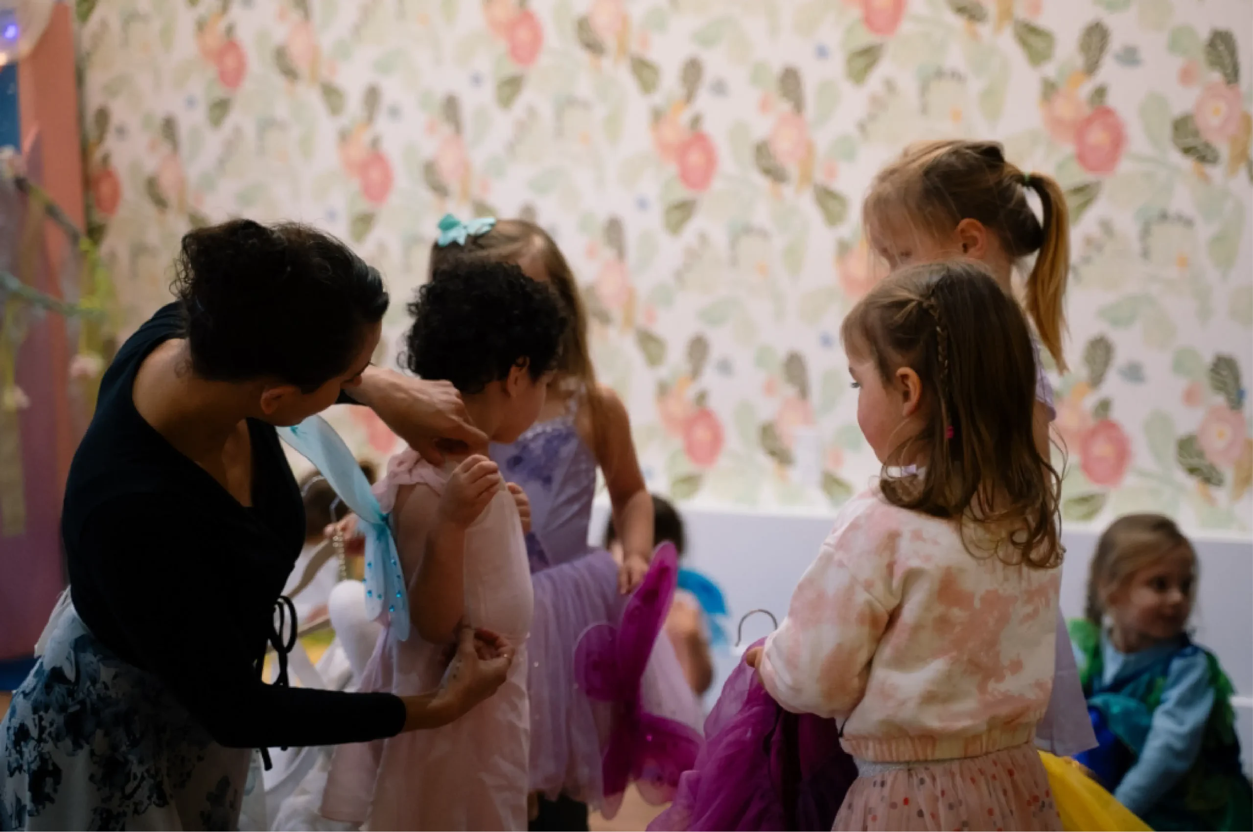 An adult helps a child adjust a dress while other children in costumes stand nearby in a room with floral wallpaper.