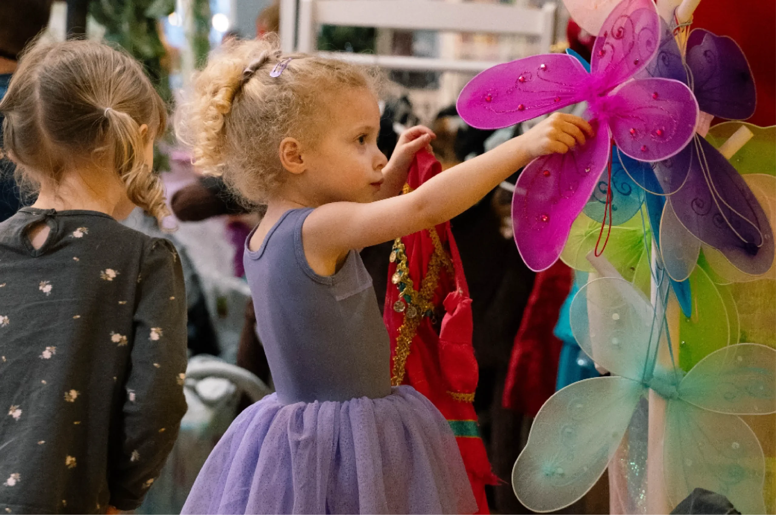 A young girl in a purple dress reaches for a pair of colorful fairy wings among various costume accessories, with another child beside her.