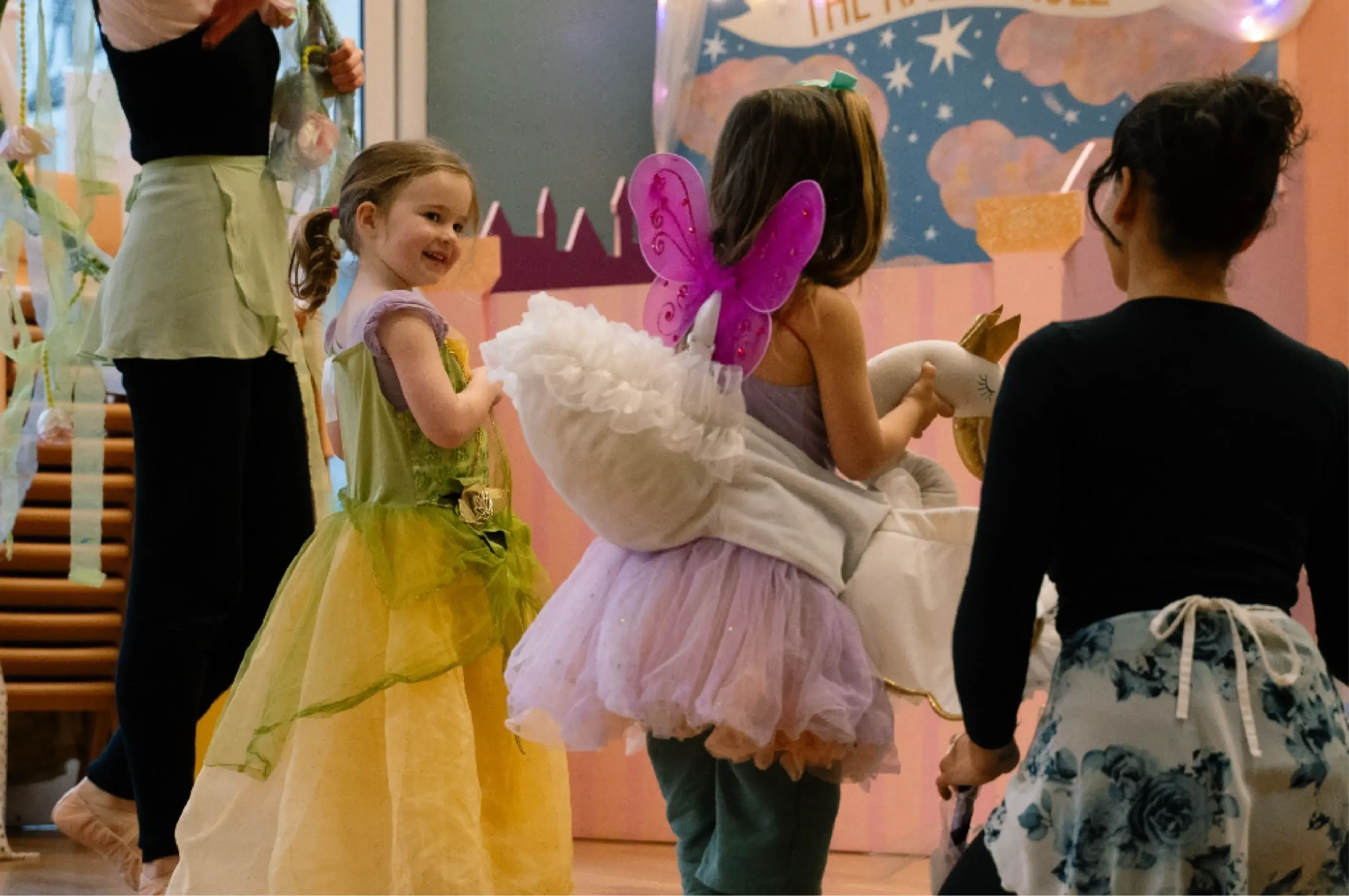 Children in colorful costumes, including a fairy and a princess, participate in an indoor party or event with pastel decorations and a painted backdrop.