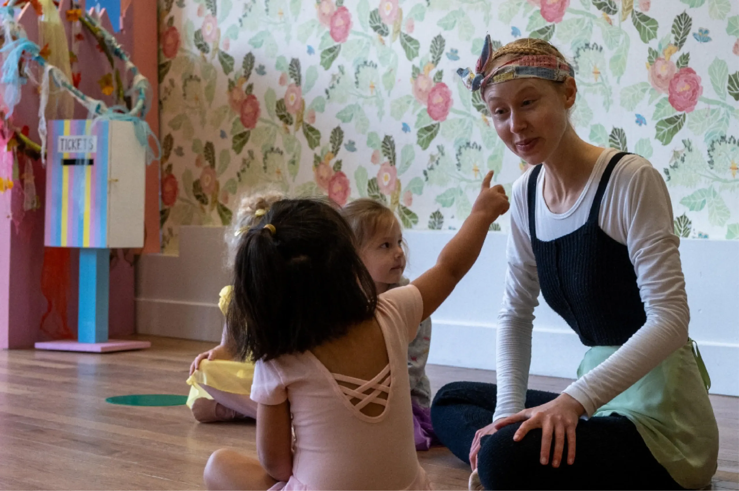 A teacher in a headscarf sits on the floor with young children in a dance studio; one child points at the teacher while talking.