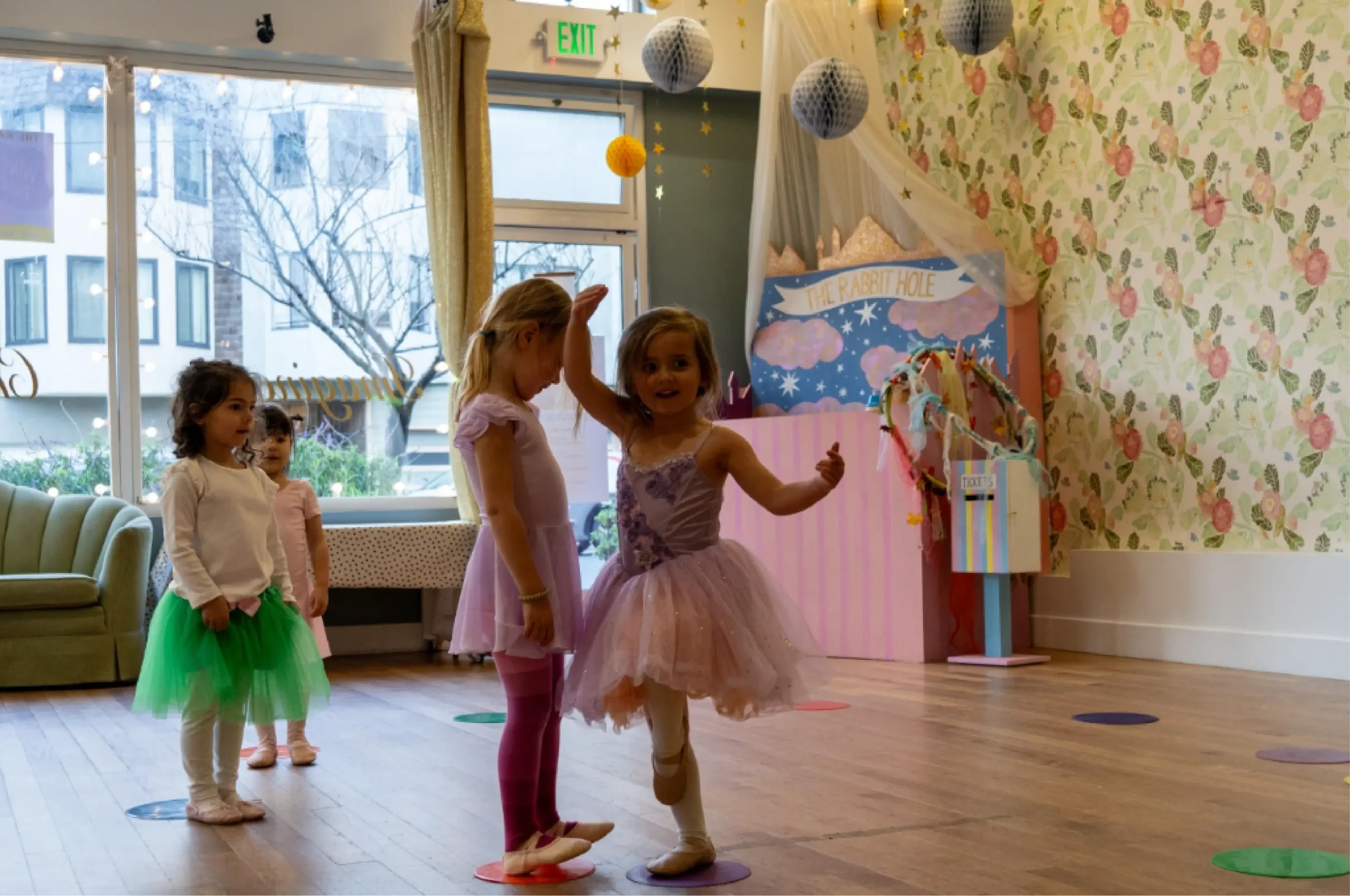 Four young children in ballet outfits stand on colored spots in a decorated dance studio with large windows and whimsical wall decor.