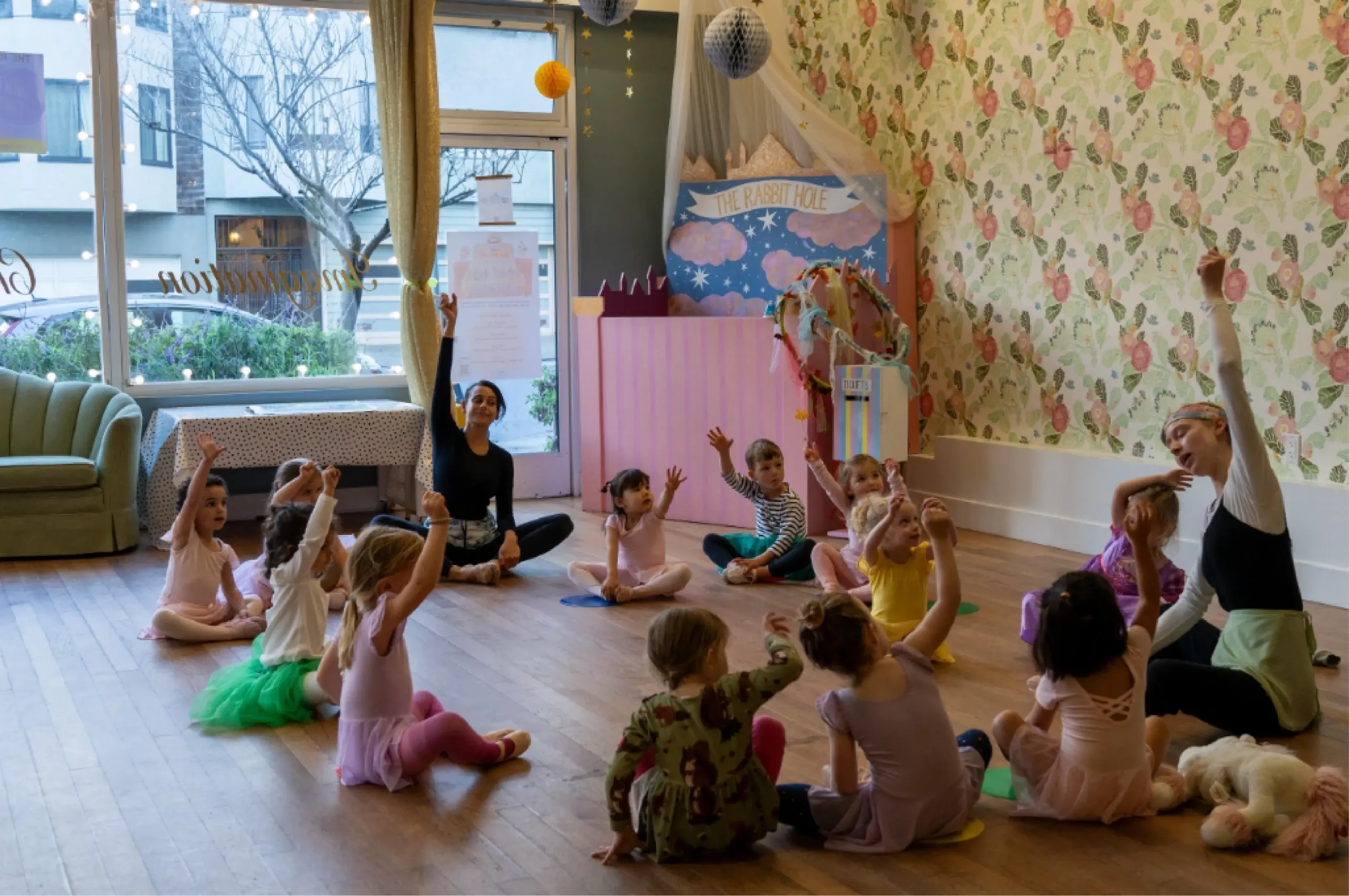 A group of young children in dance attire sit in a circle with two instructors in a decorated studio, raising their hands during a class activity.