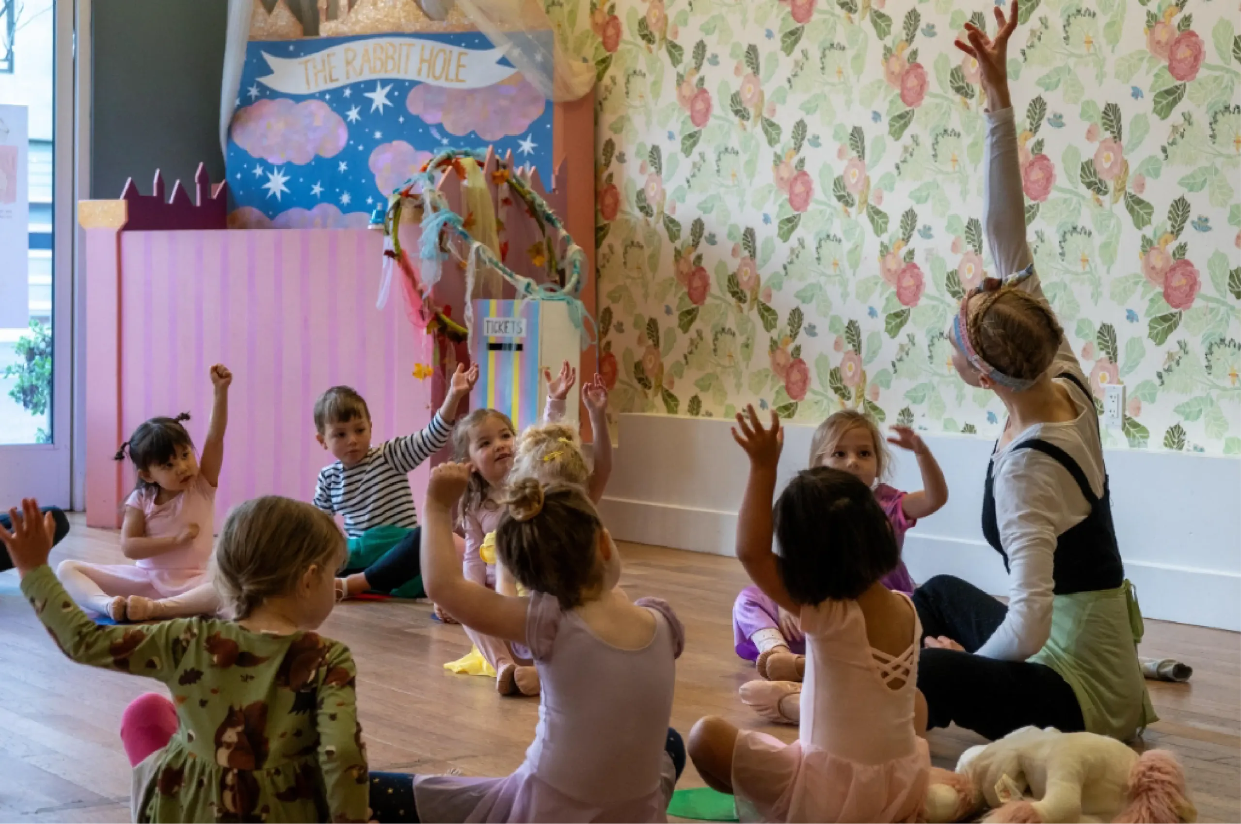 A group of young children sit on the floor with their hands raised, participating in an activity led by an adult in a decorated room with floral wallpaper and a "The Rabbit Hole" sign.
