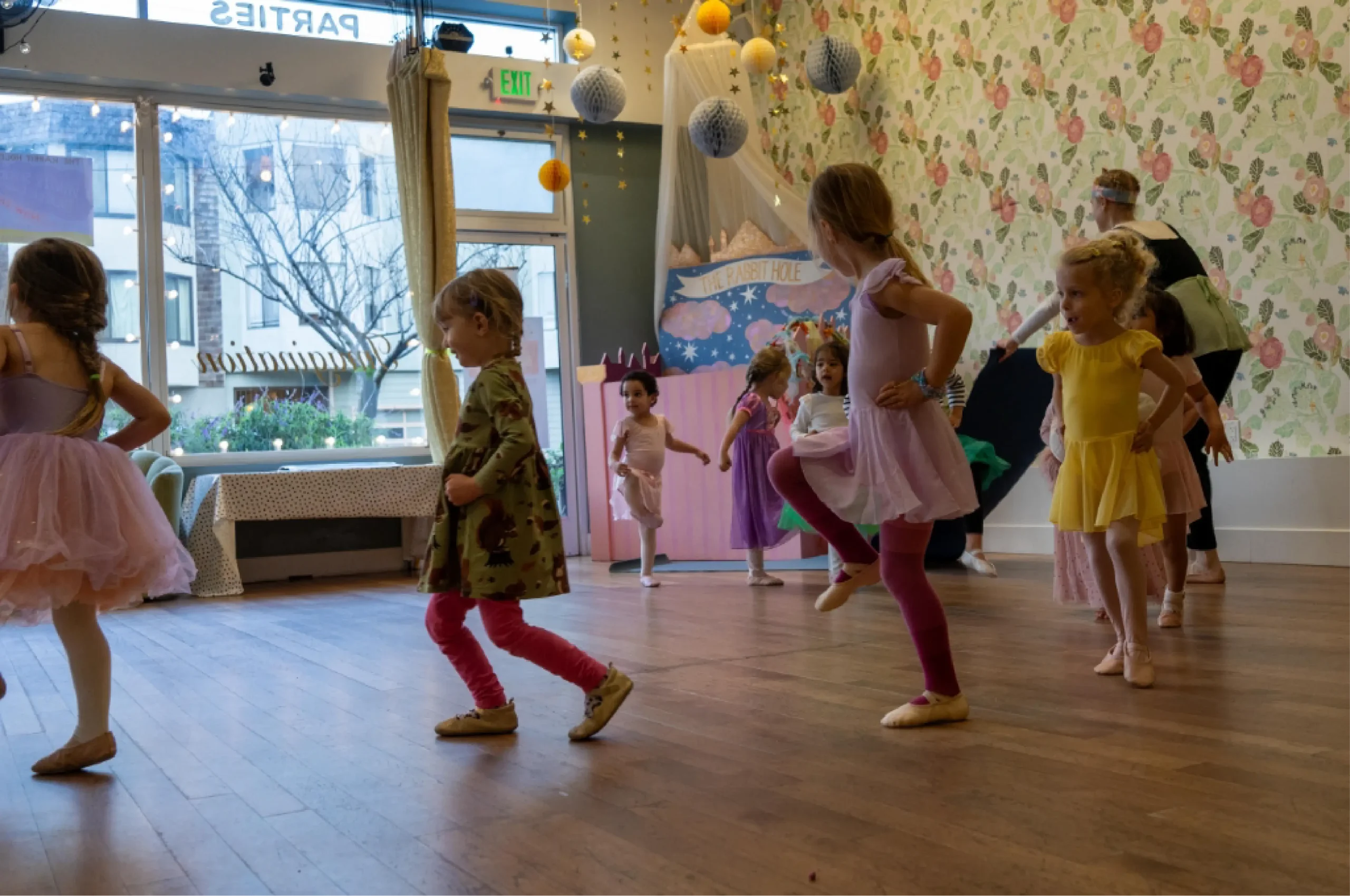 Young children in colorful costumes participate in a dance class in a decorated studio with a floral wallpaper and wooden floor.