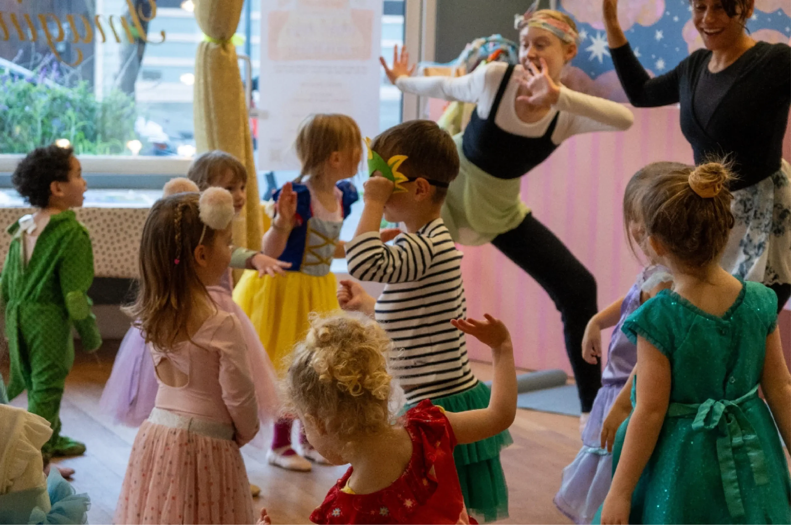 A group of young children in costumes participate in a dance activity, led by two adults in a decorated indoor space.