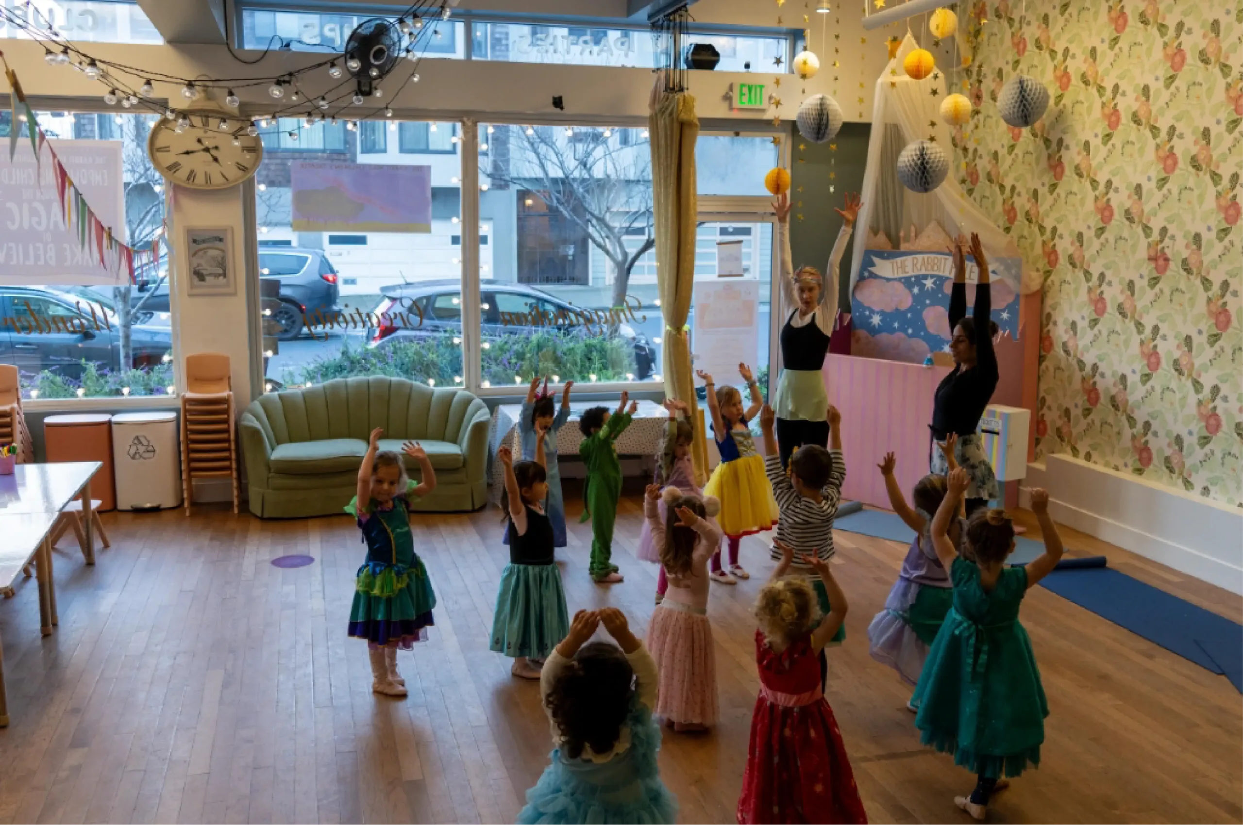 A group of young children in colorful costumes participate in a dance class led by two adult instructors in a decorated studio with large windows.