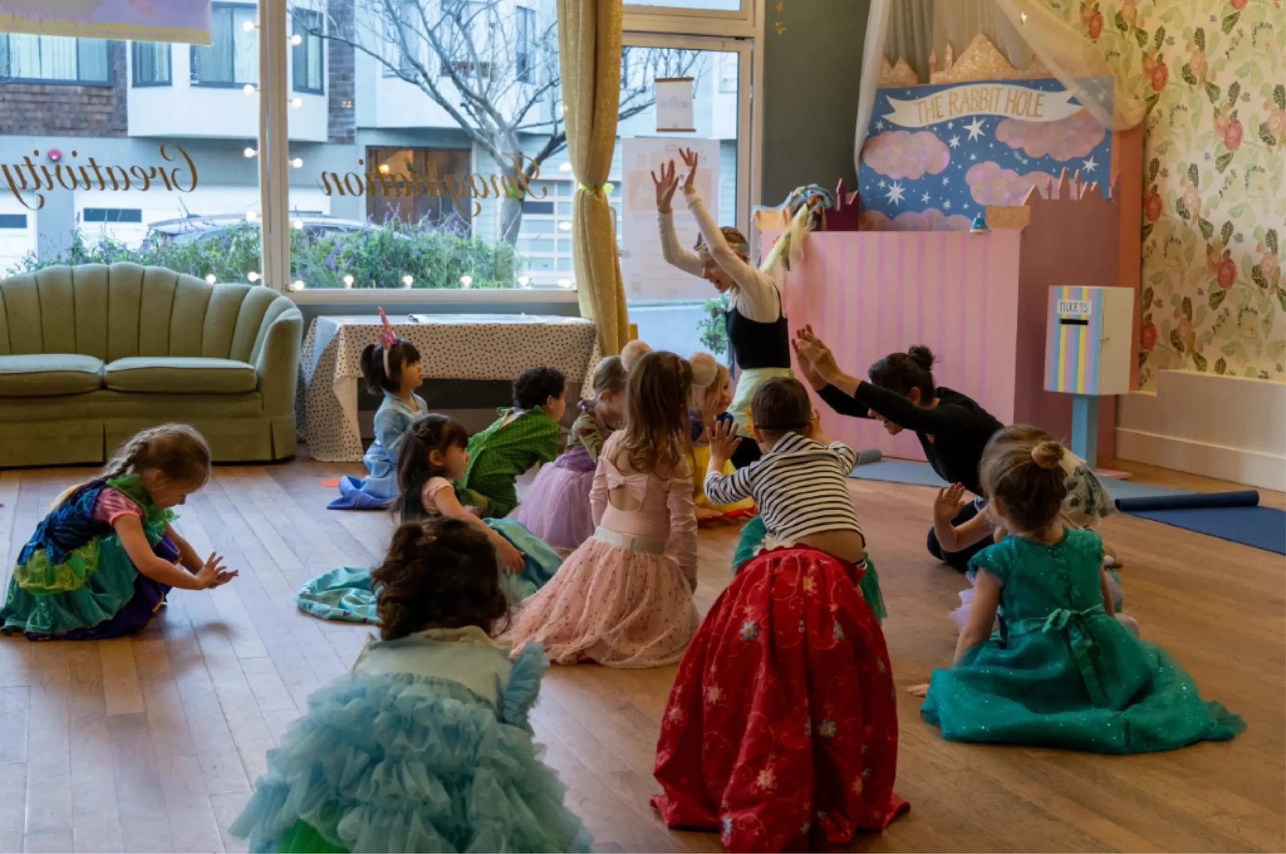 A group of young children in costumes sit and raise their arms in a circle on a wooden floor, guided by an adult in a decorated room with large windows.