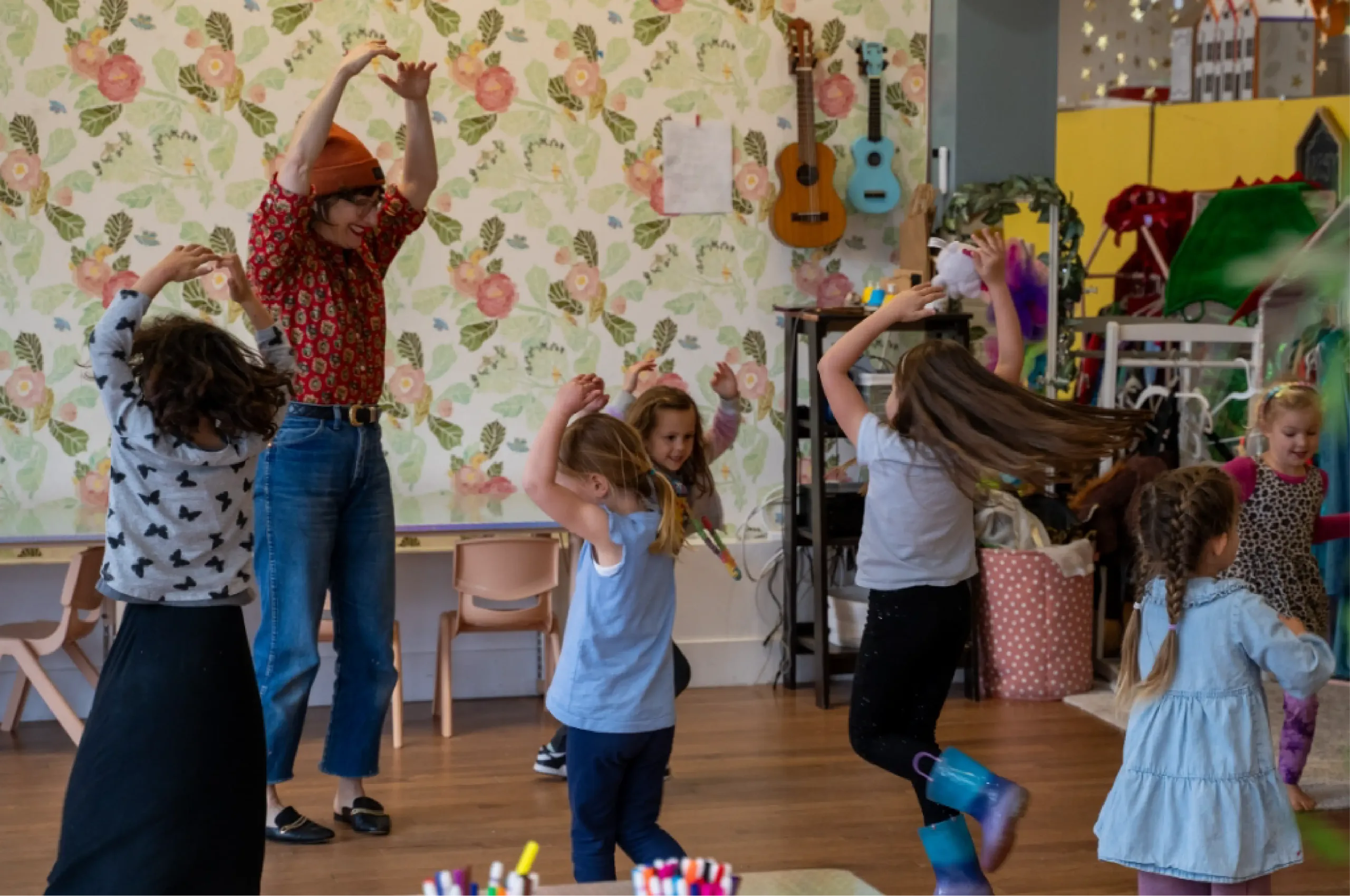 A group of children and an adult dance in a colorful room with floral wallpaper, musical instruments, and craft supplies visible in the background.