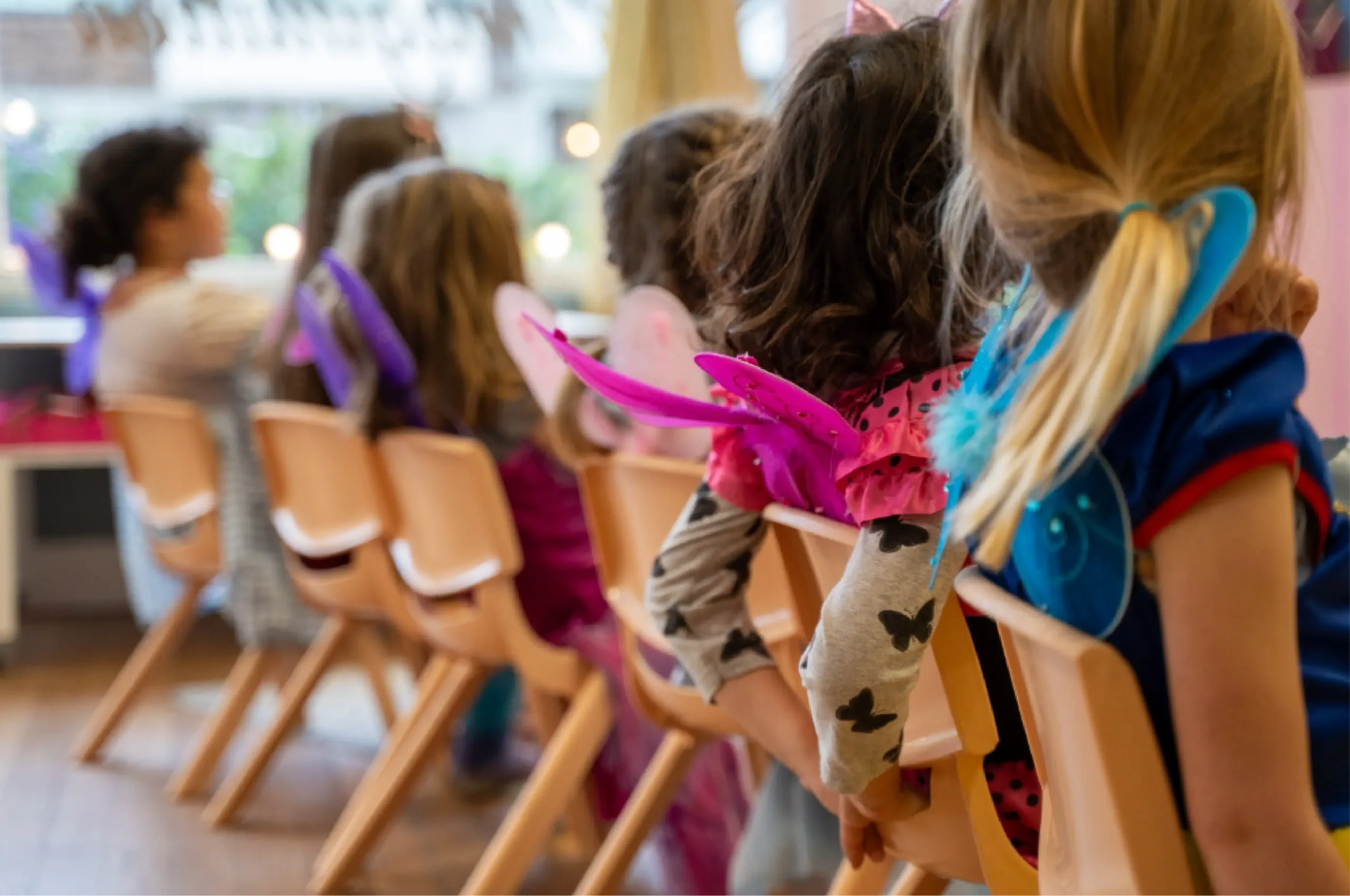 Children dressed in colorful costumes with wings sit in a row of chairs indoors, facing away from the camera.