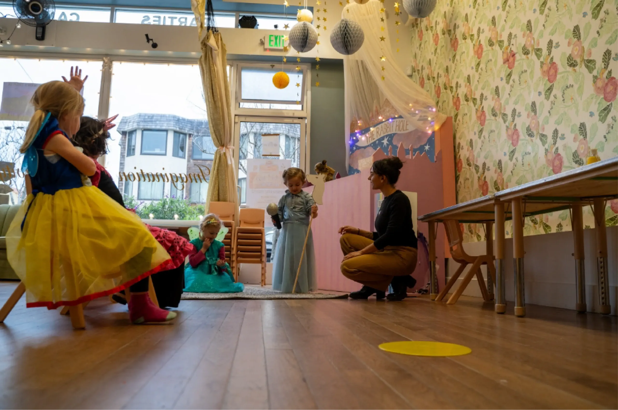 Several young children in costumes sit and watch as one child stands in front of an adult in a decorated room with floral wallpaper and string lights.