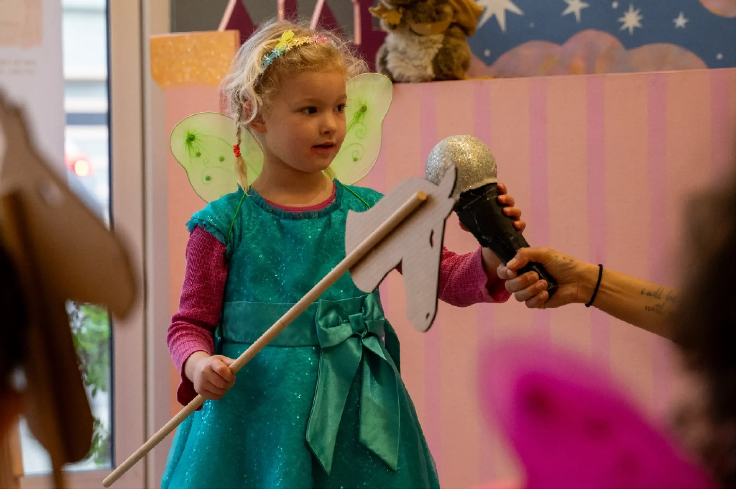 A young child in a fairy costume holds a stick horse and stands in front of a toy microphone held by an adult.