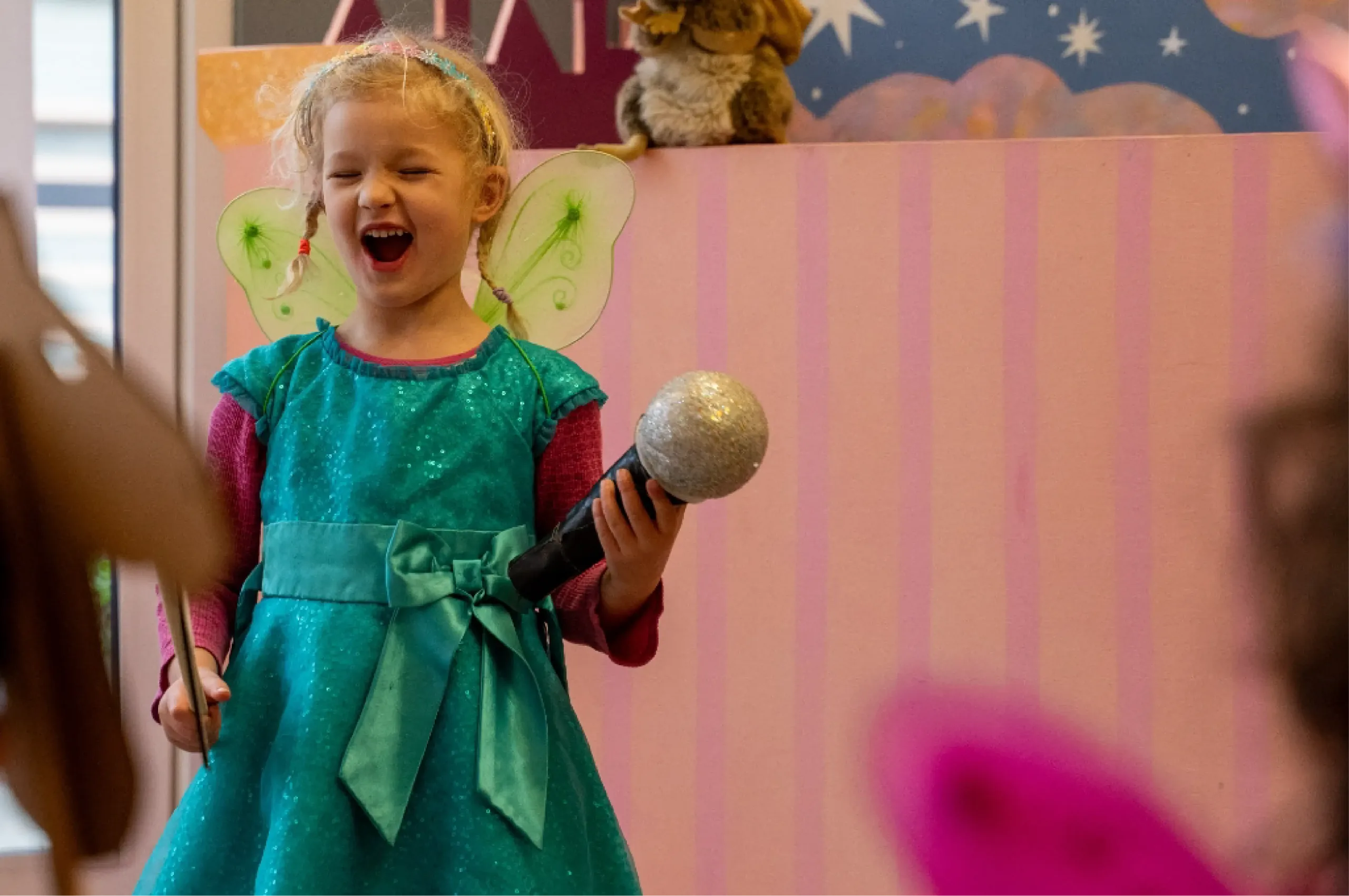Young girl in a turquoise fairy costume holds a toy microphone and smiles with eyes closed, standing in front of a pink striped background.