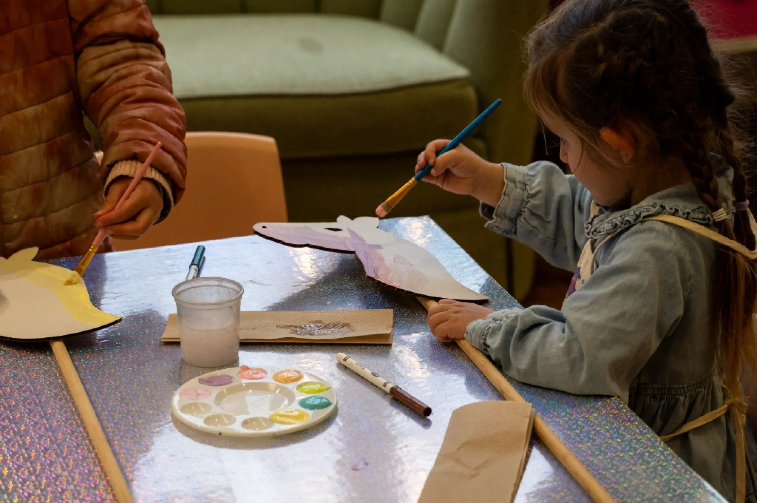 Two children sit at a table painting paper cutouts with brushes and watercolors; art supplies and napkins are scattered on the shiny tabletop.