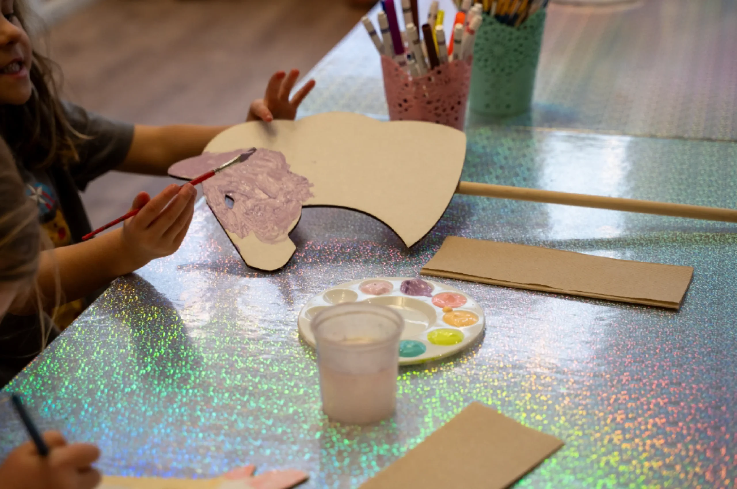 Child painting a cardboard cutout with light purple paint at a table, with paint palette, water cup, brushes, and brown paper nearby.