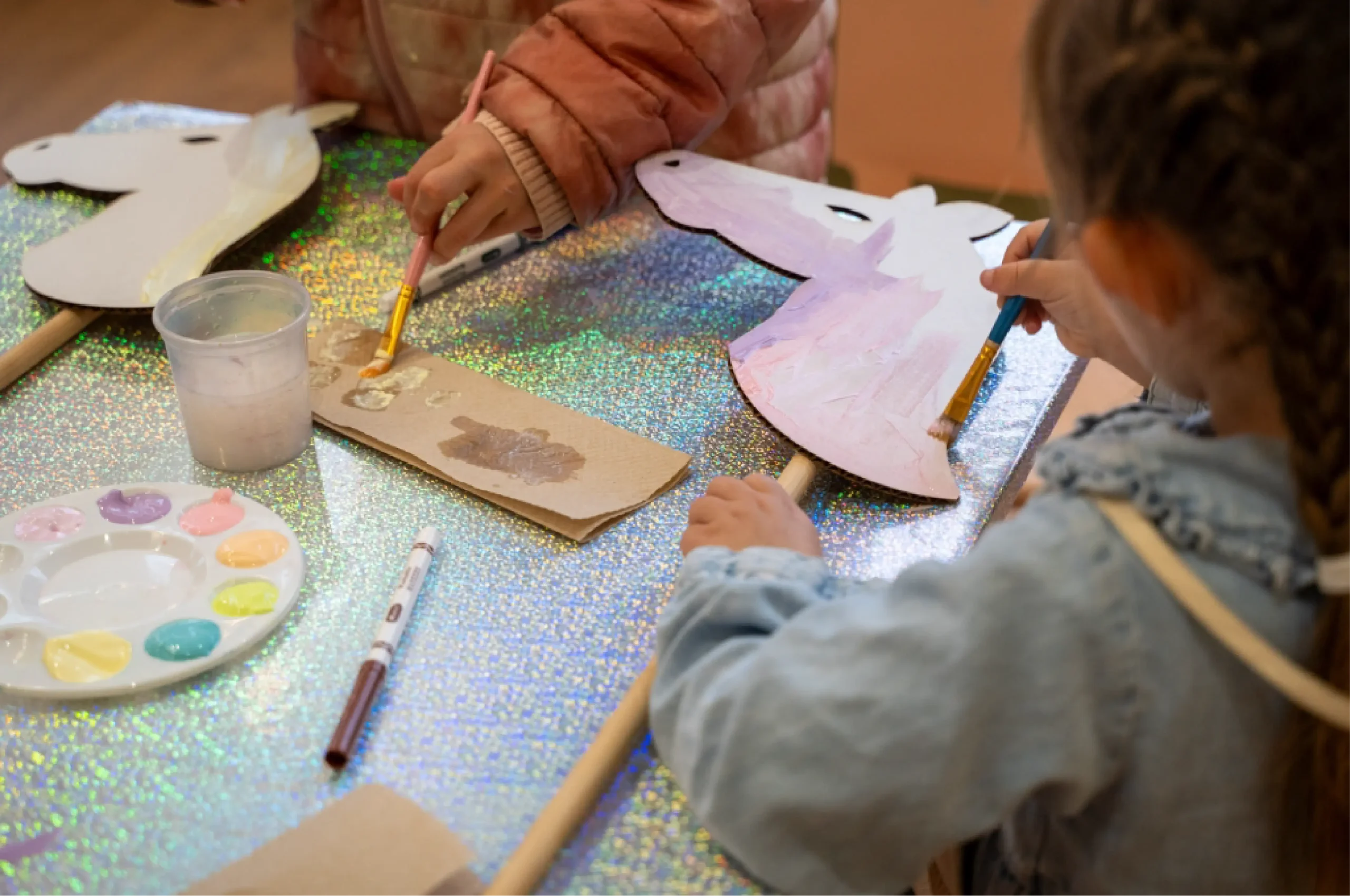 Two children paint unicorn masks and cardboard pieces at a table covered with iridescent material; paint supplies and brushes are visible.