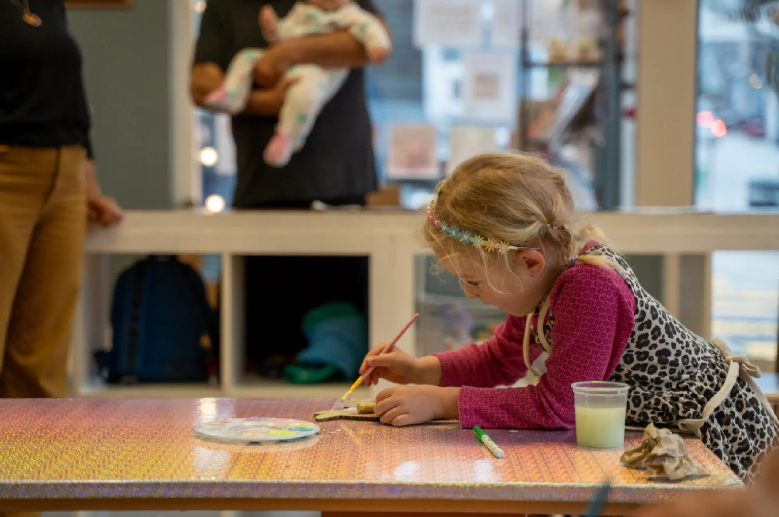A young girl sits at a table painting with a brush, while two adults, one holding a baby, stand in the background.