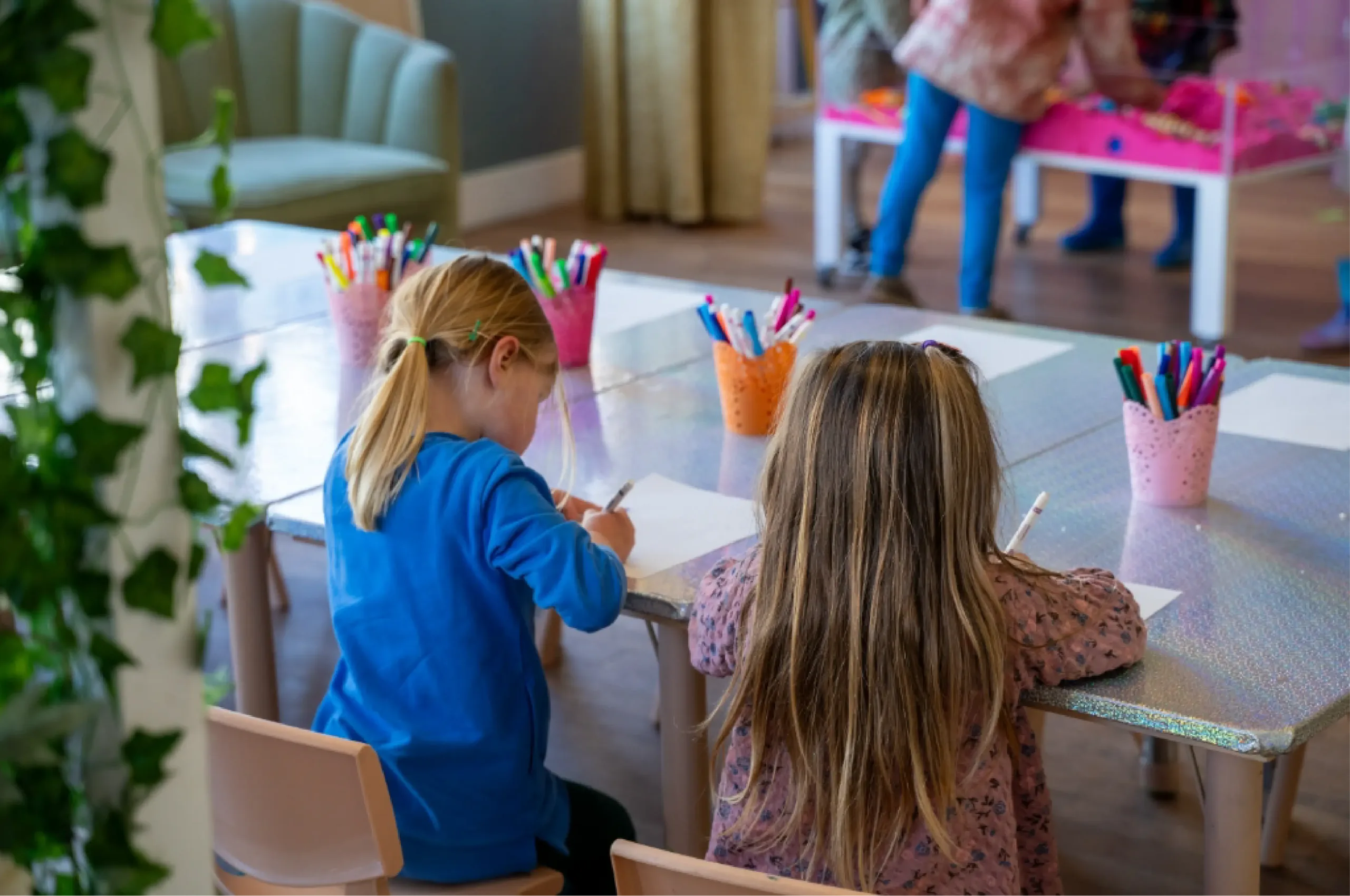 Two children sit at a table drawing with colored markers in a classroom or playroom setting. Other children are seen standing and playing in the background.