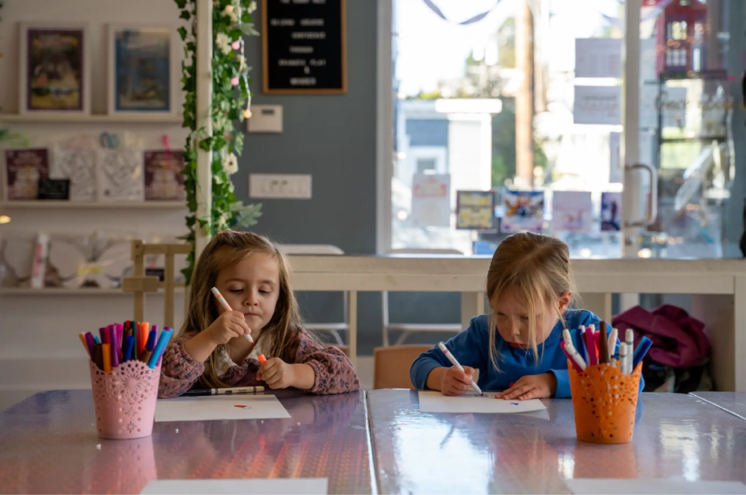 Two young children sit at a table in a bright room, drawing on paper with markers. Art supplies are visible on the table.