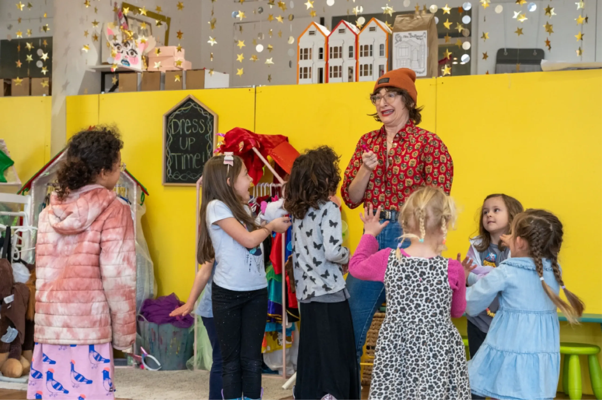 A woman stands smiling and talking with a group of young children in a colorful playroom decorated with costumes and star decorations.