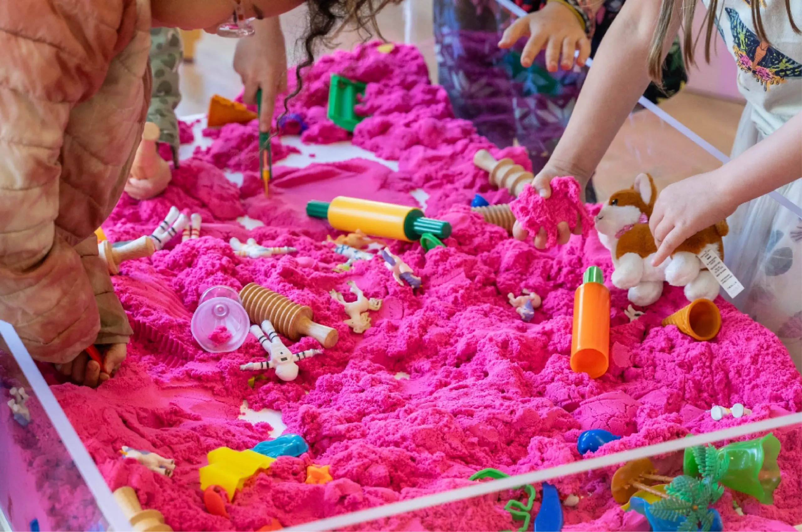 Children’s hands playing with bright pink kinetic sand, toy rolling pins, animal figurines, and plastic molds on a tabletop sensory activity setup.