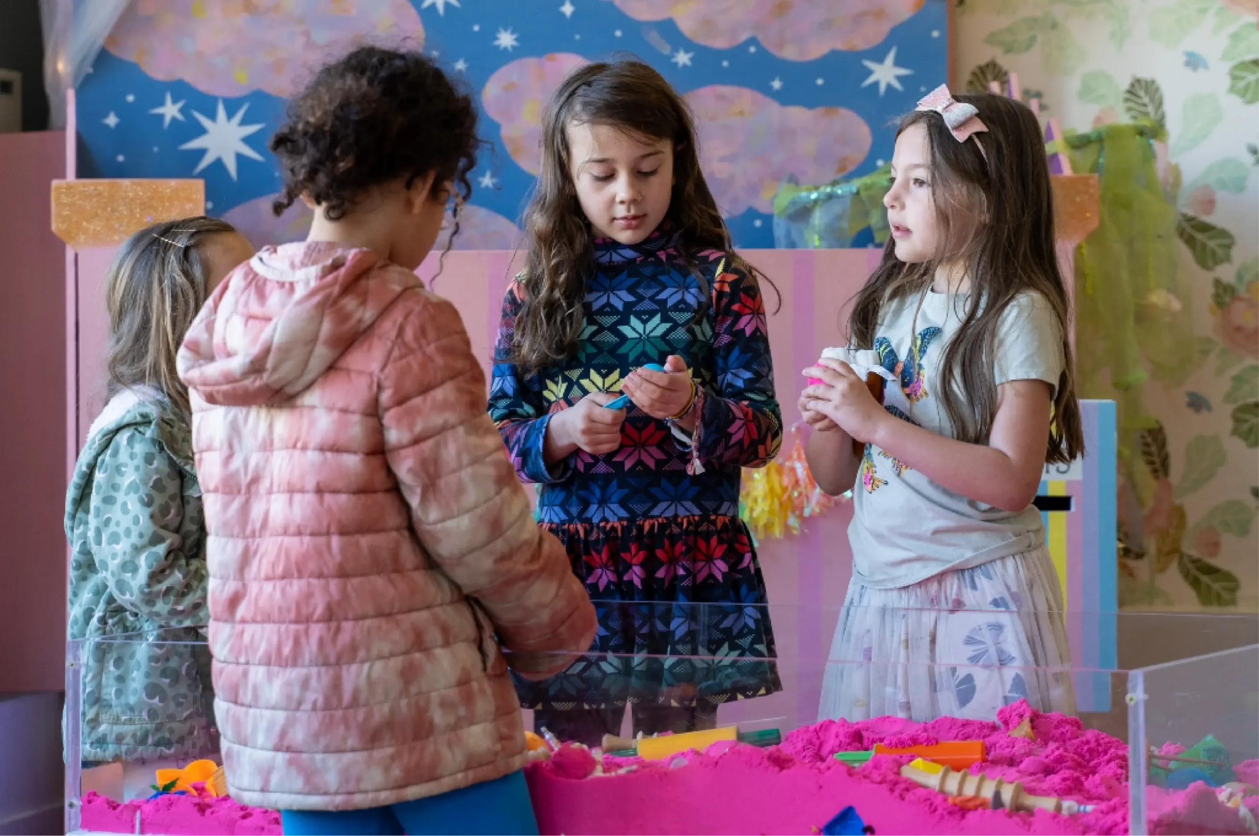 Four young girls stand around a table filled with bright pink sand, playing and talking together in a colorful, decorated room.