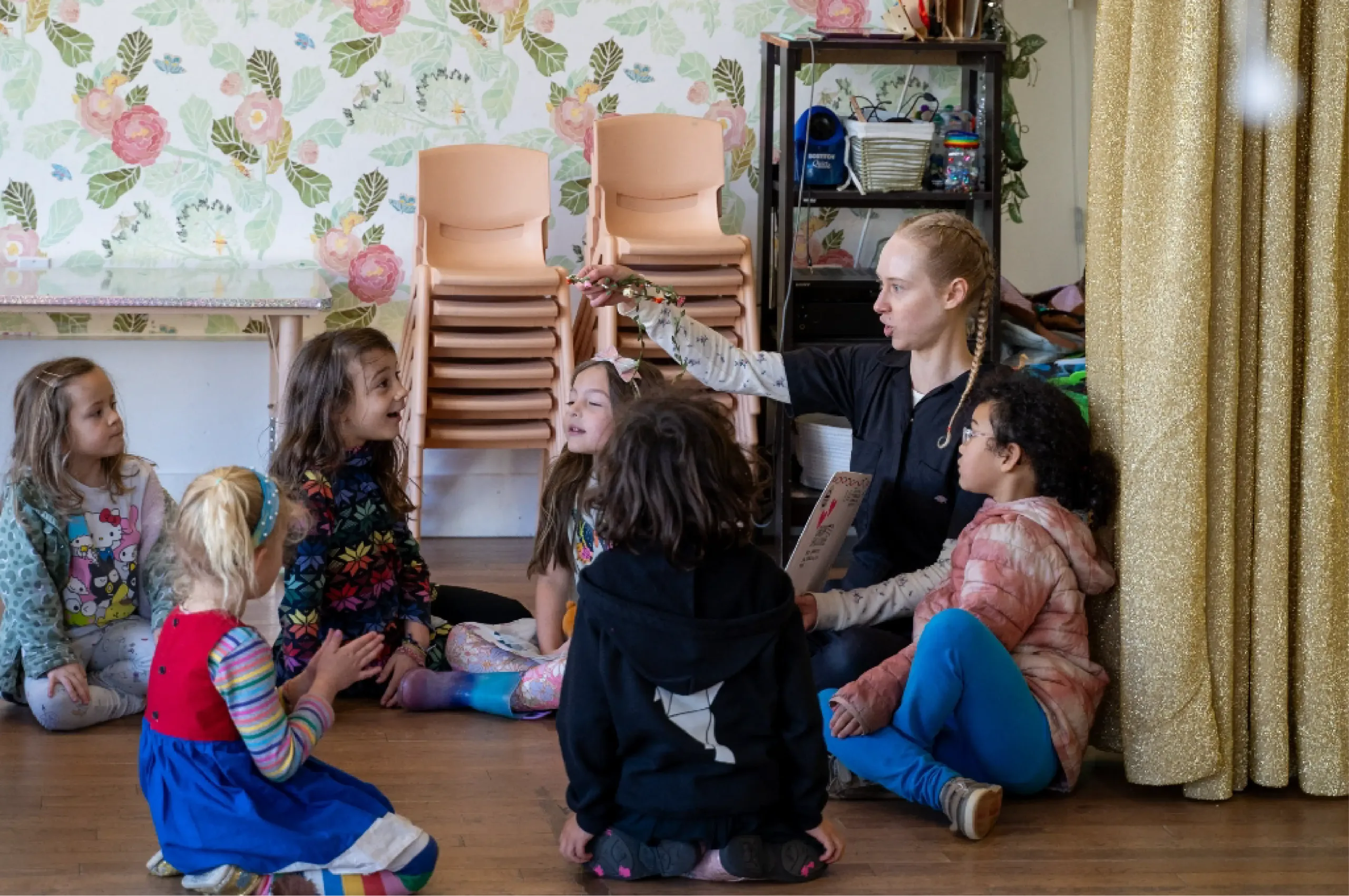 An adult sits on the floor showing an object to a group of young children who are seated in a circle in a decorated room.