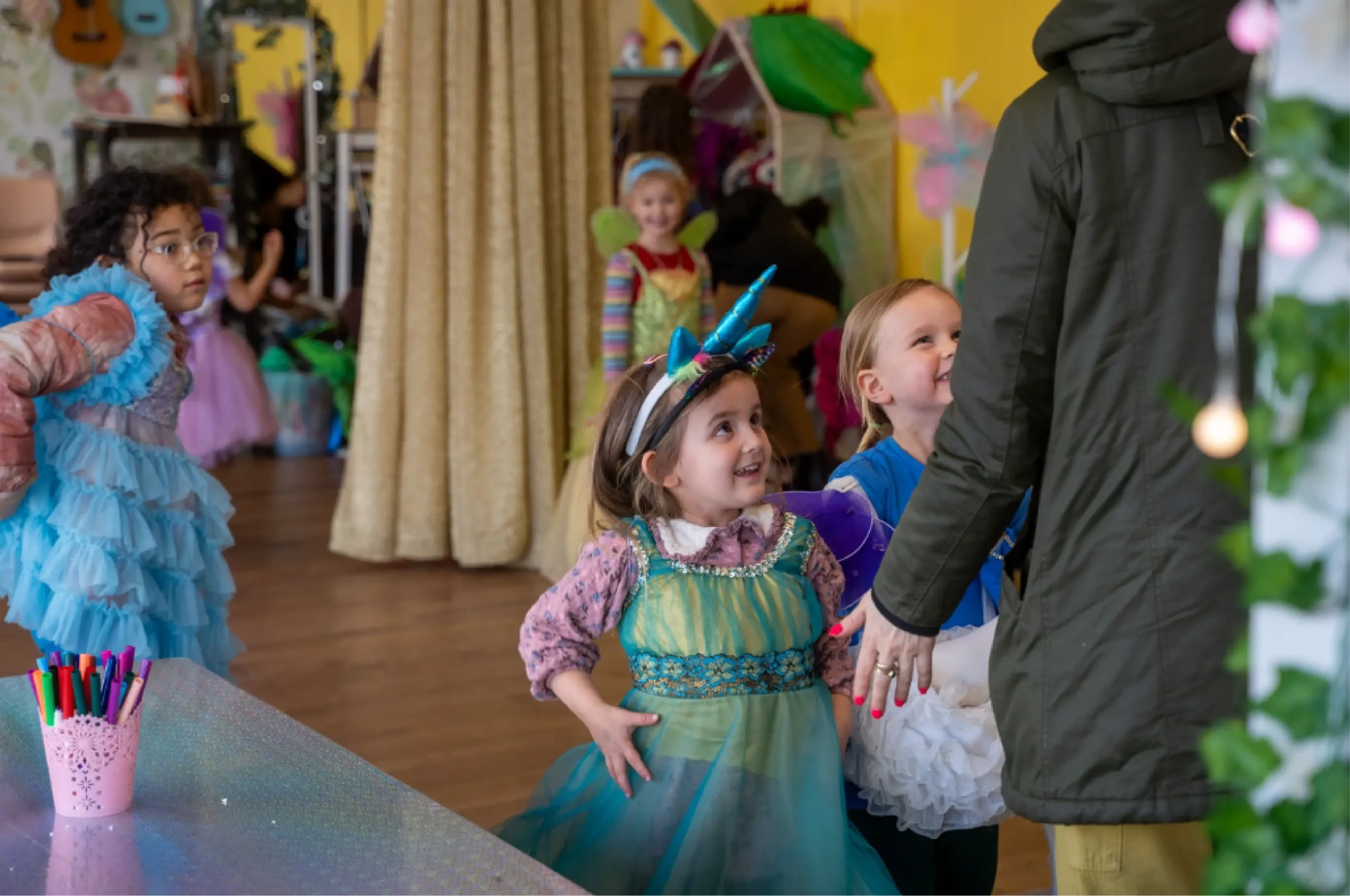 Children in colorful costumes stand in a line indoors, with a woman in a dark coat at the front. Art supplies and decorations are visible in the background.