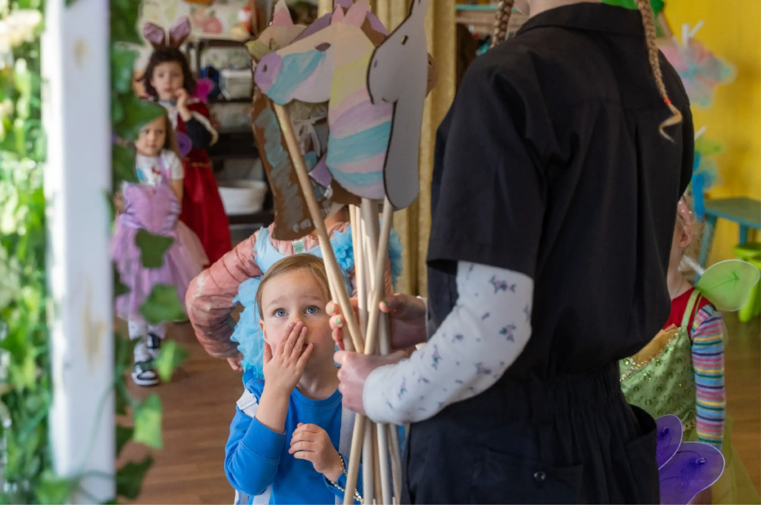 A young child in blue looks up and covers their mouth while an adult holds a bunch of unicorn hobby horses in a colorful indoor setting with other children in the background.