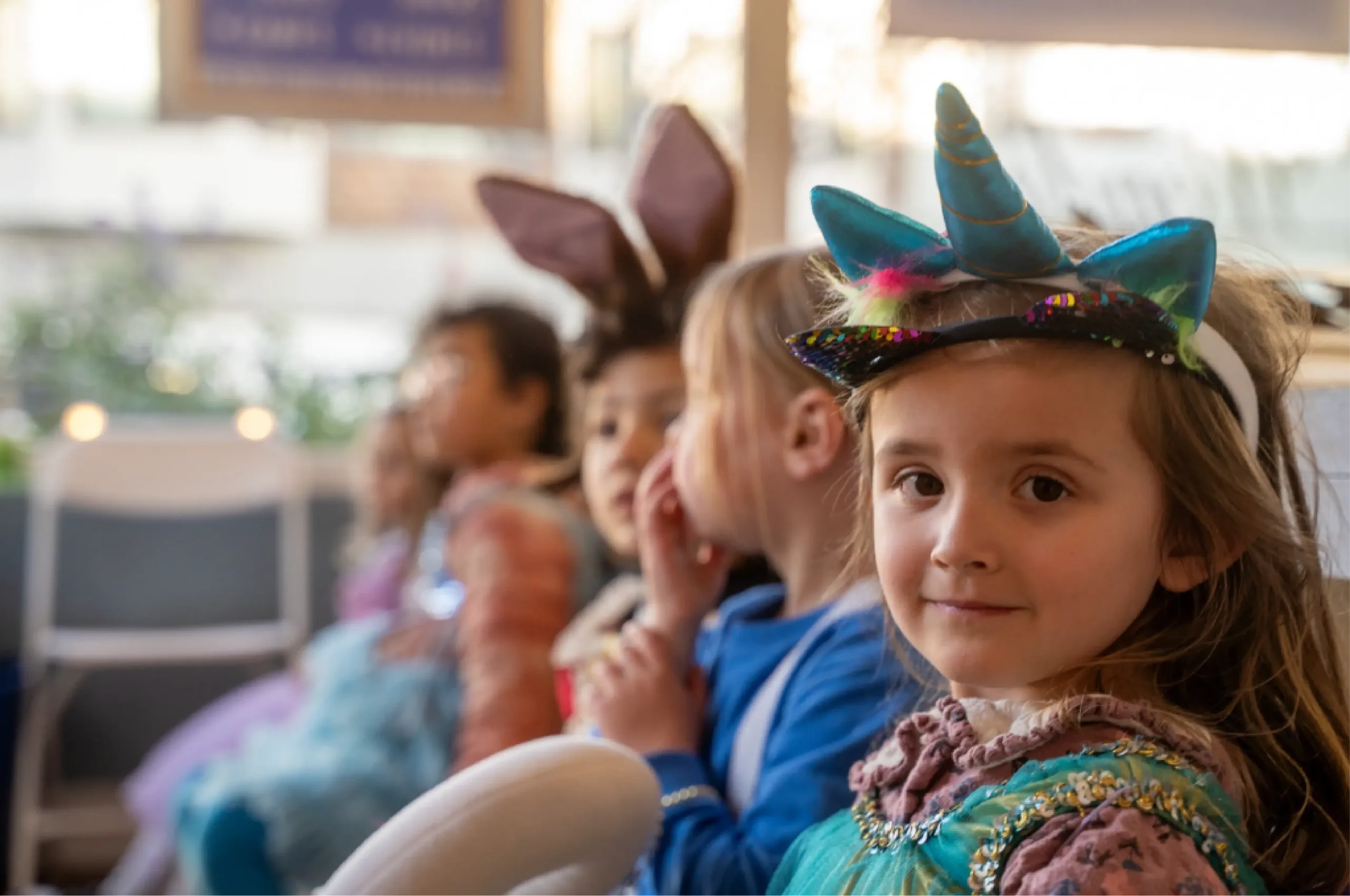 Children sit in a row wearing animal and fantasy-themed costumes, including a unicorn horn headband and bunny ears, at what appears to be a party or event.