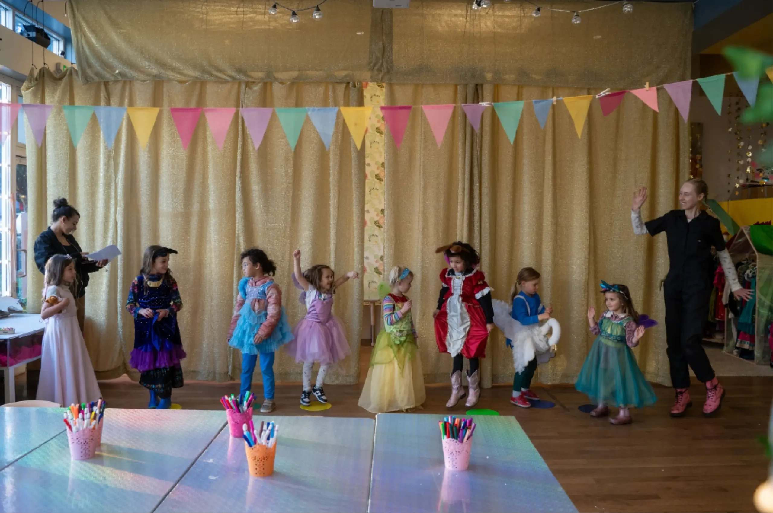 A group of children in costumes stand in a line with two adults in a decorated room, colorful bunting overhead and craft supplies on tables in the foreground.
