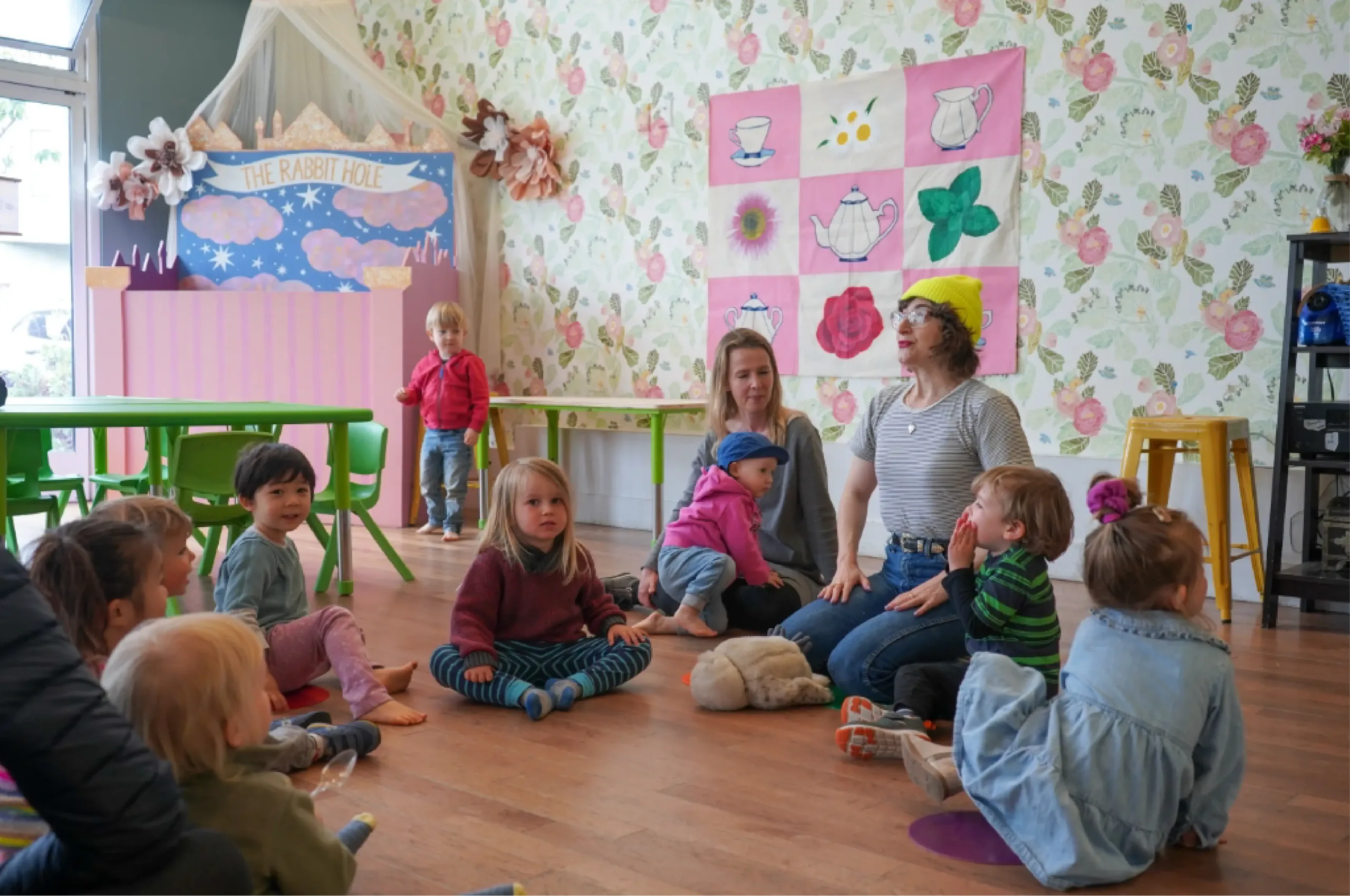 Children and two adults sit in a circle on a classroom floor decorated with floral wallpaper, a pink banner, and wall art featuring teapots and flowers.