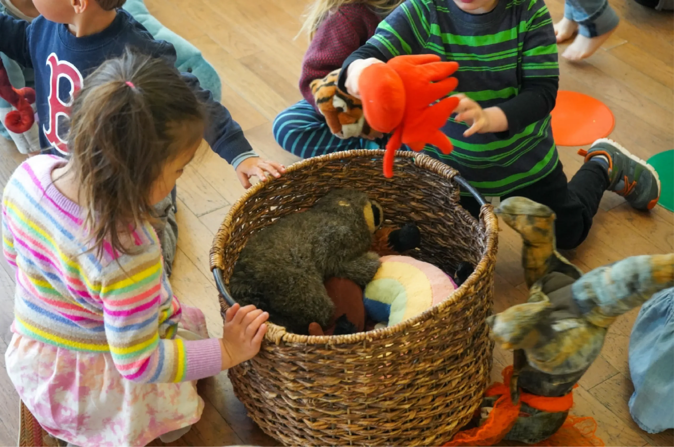 Several young children sit on a wooden floor around a wicker basket filled with stuffed animals and puppets, reaching in and playing with the toys.