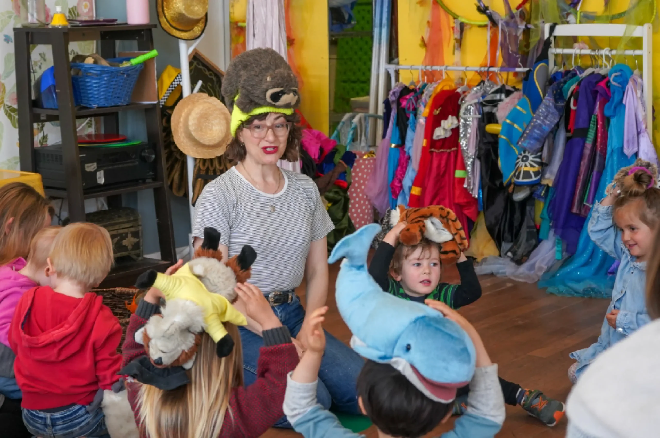 A woman wearing a bear hat sits with young children in a playroom; some children wear animal hats, and costumes hang on racks in the background.
