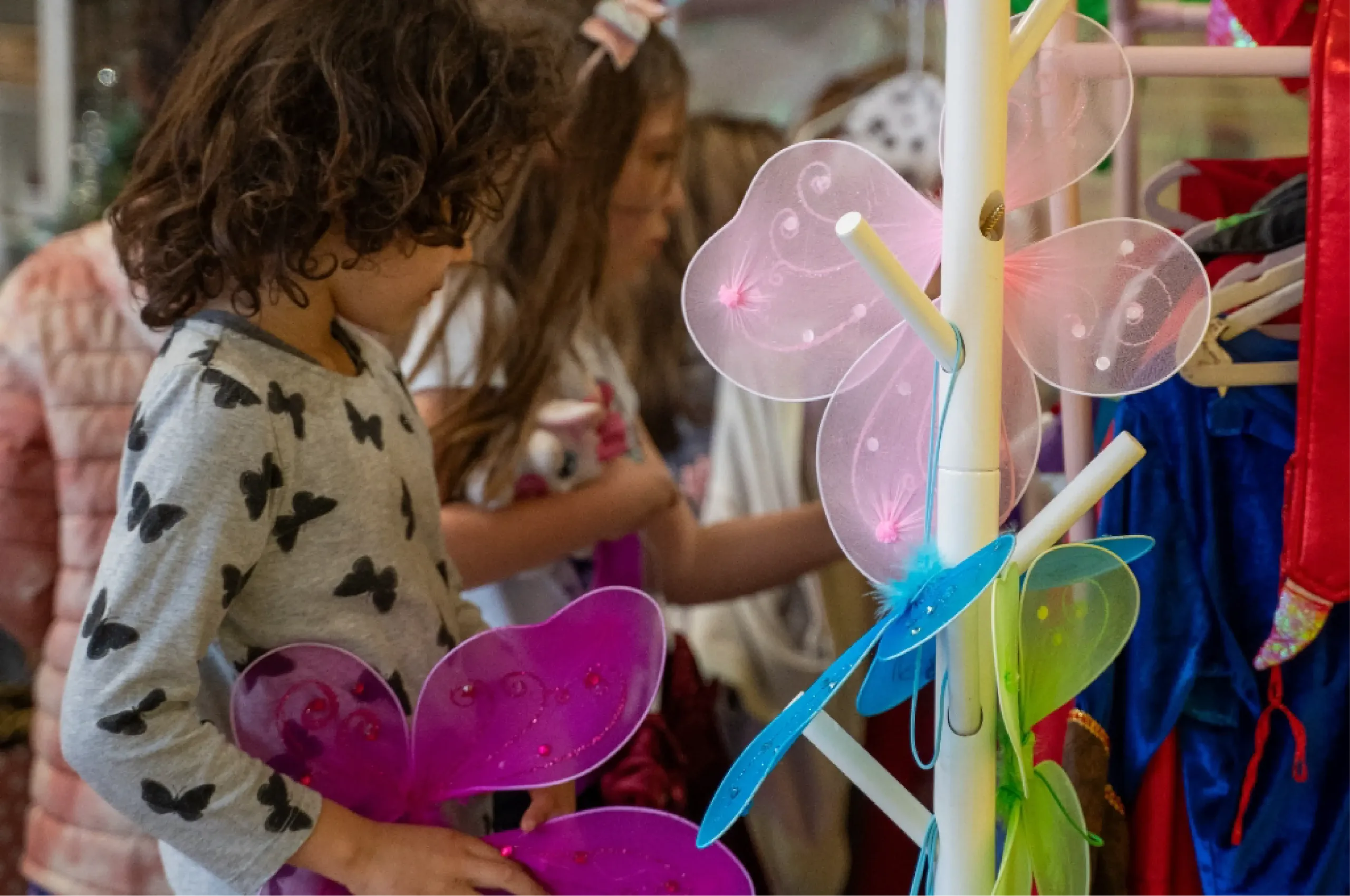 Children select colorful fairy wings from a display rack in a costume shop.