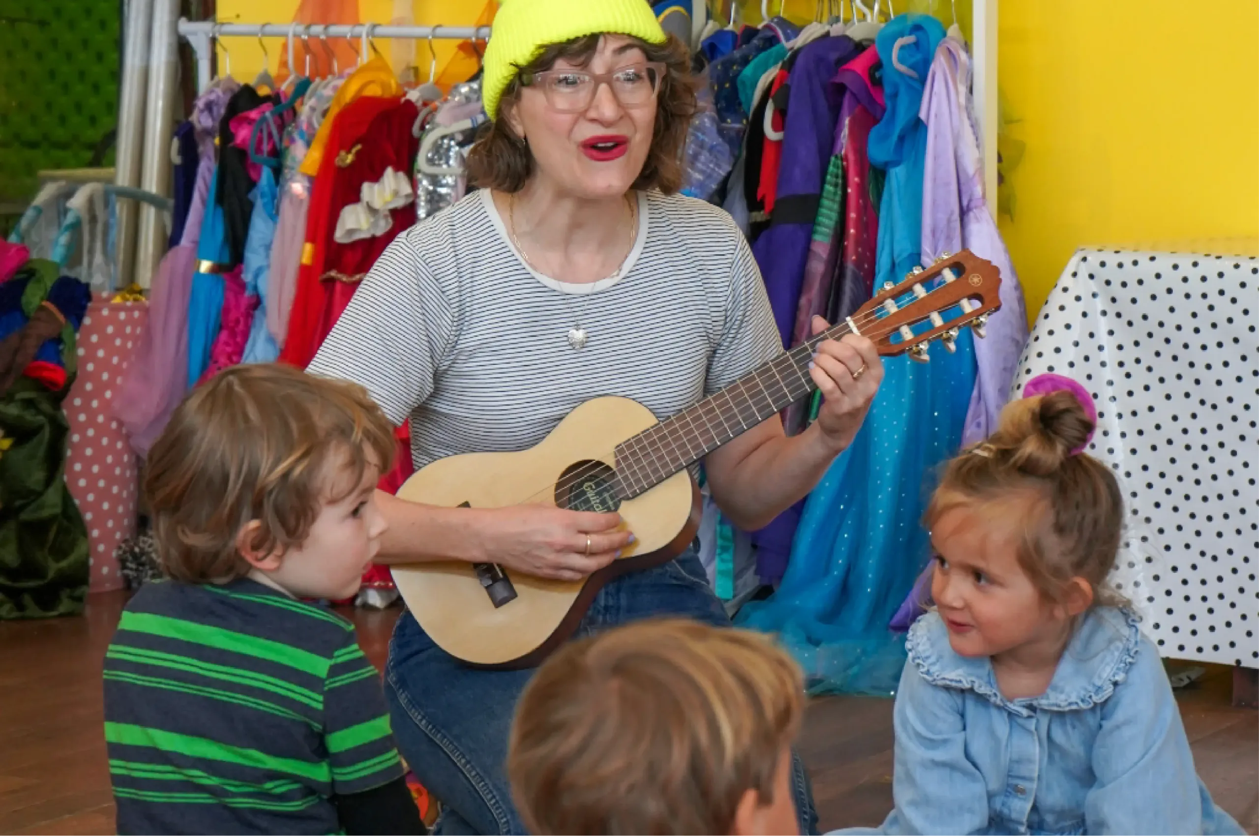 An adult wearing a yellow hat plays a small guitar while three young children sit and listen; colorful costumes hang in the background.