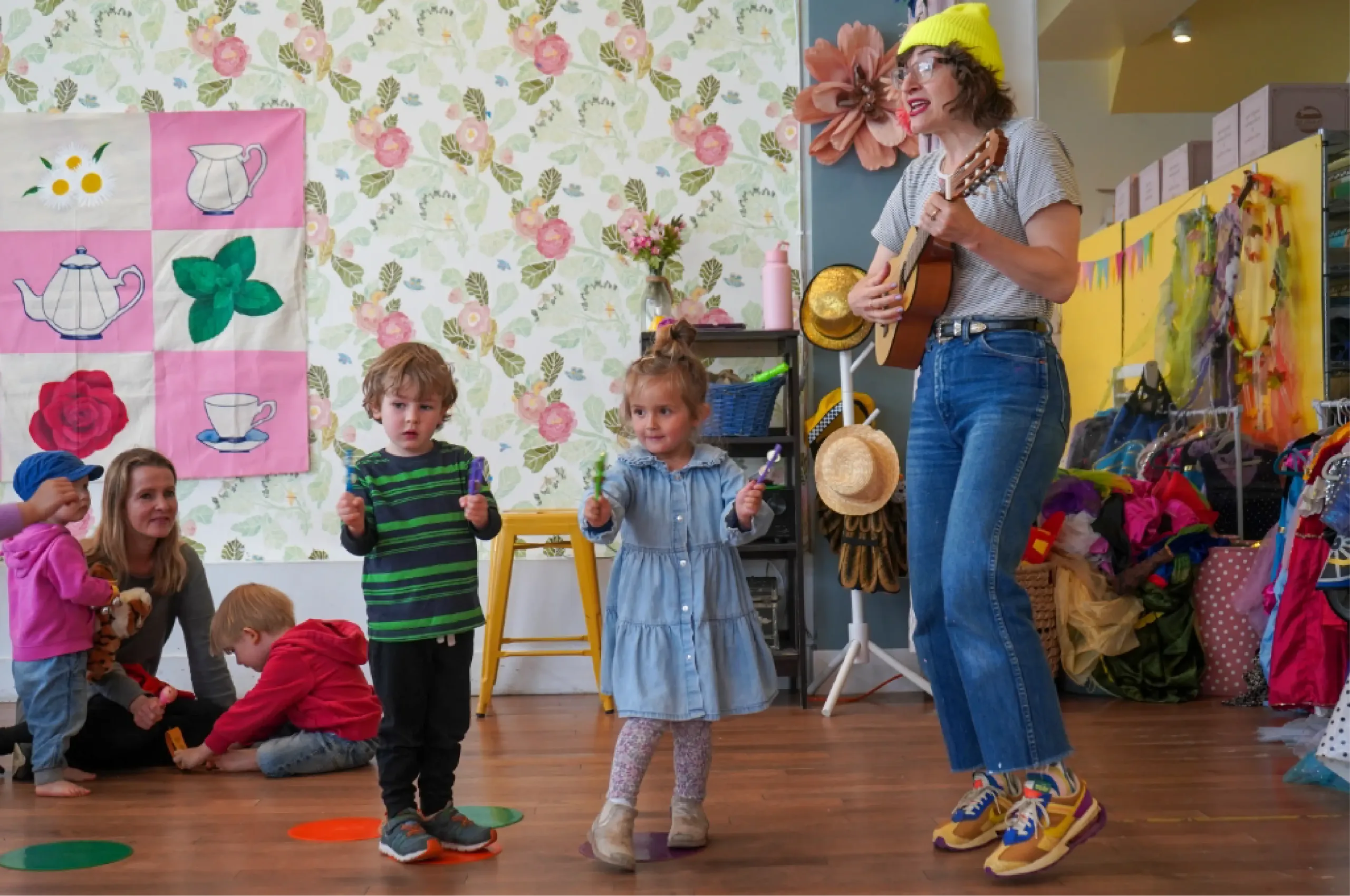 A woman plays a ukulele while two young children stand and gesture; other children play in the background of a colorful, decorated room.