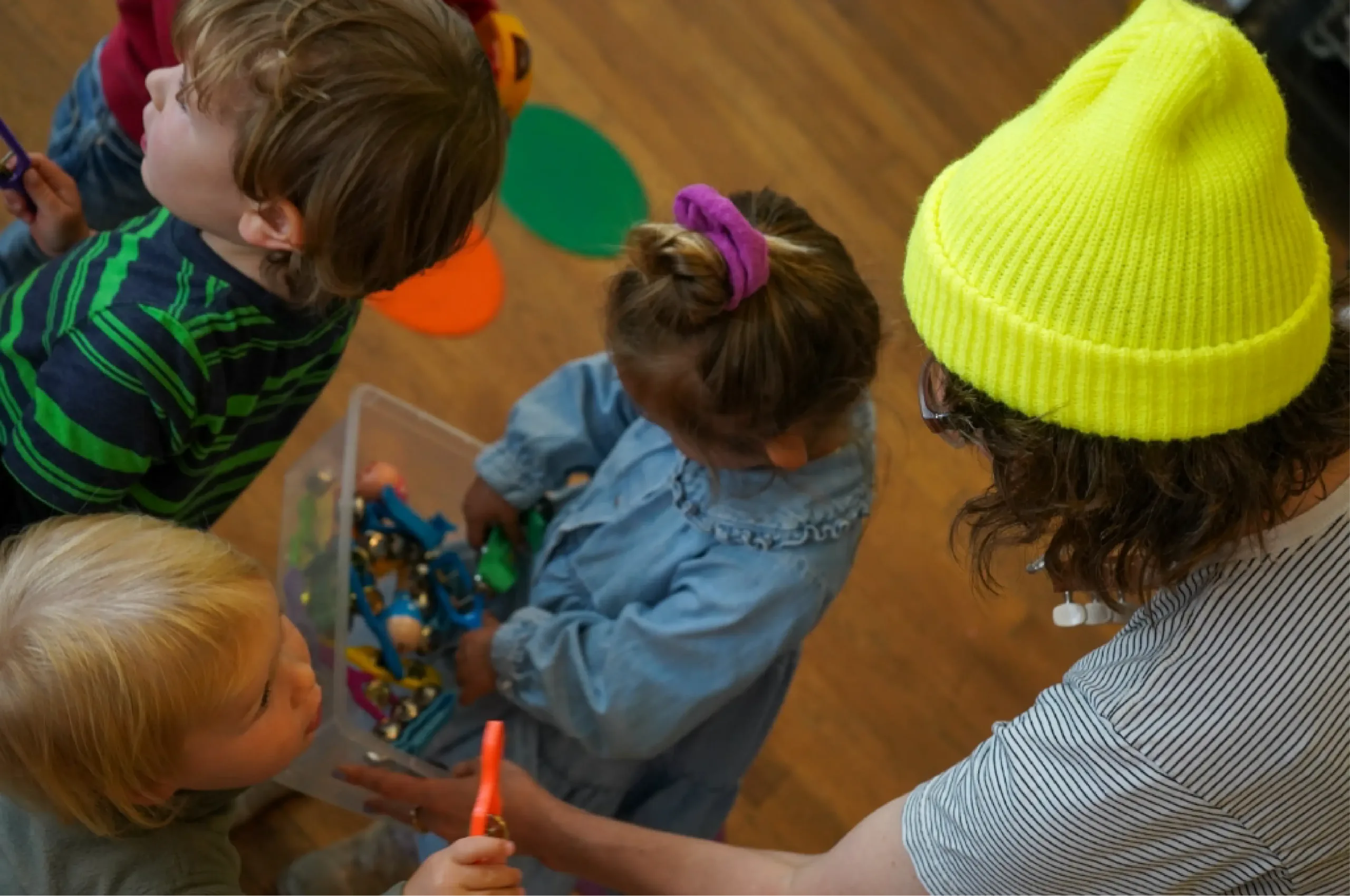 Four young children and an adult gather around a plastic container filled with toys on a wooden floor, viewed from above.
