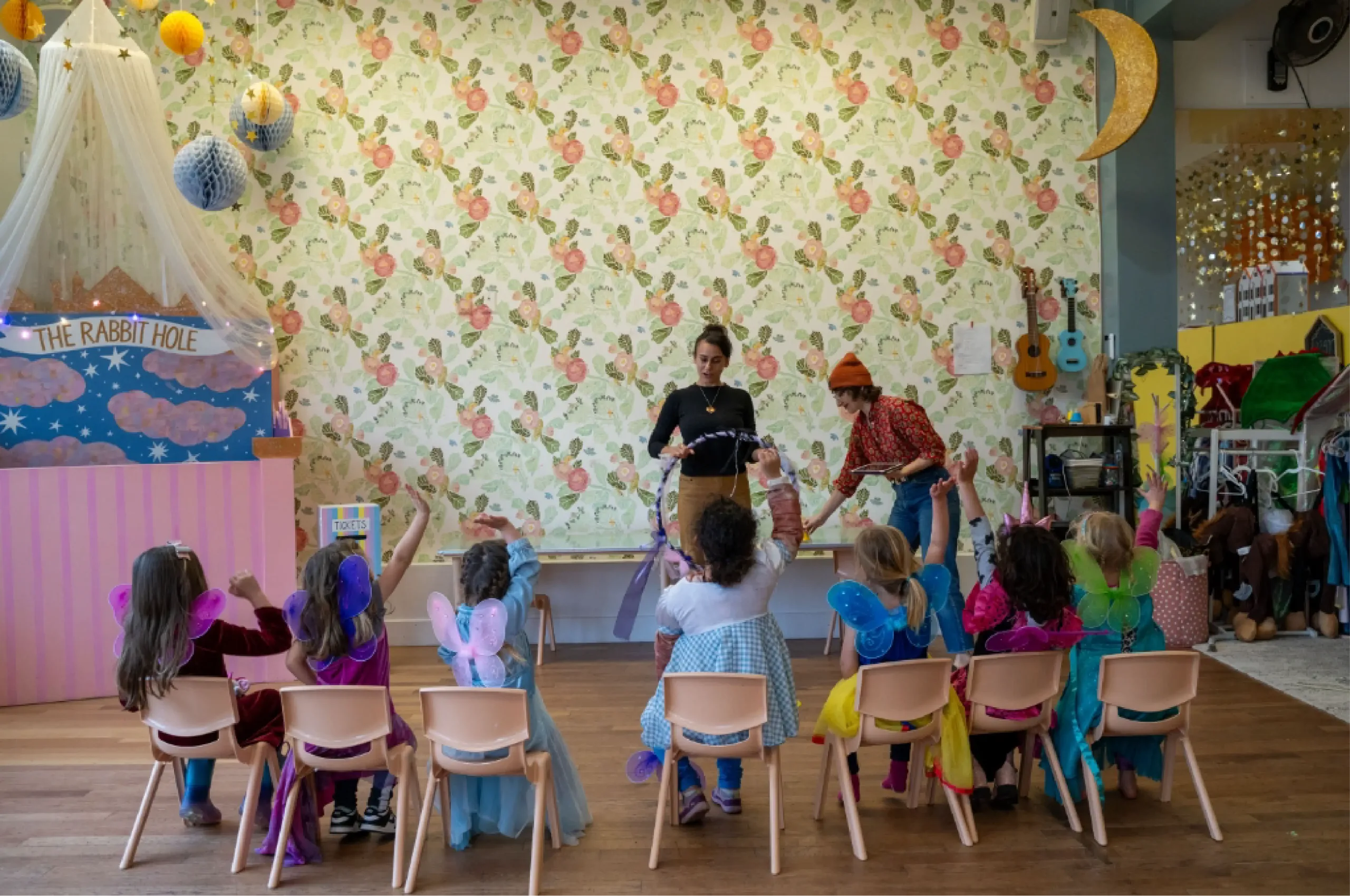 Children in costumes sit on chairs facing two adults leading an activity in a decorated classroom with floral wallpaper and whimsical decor.