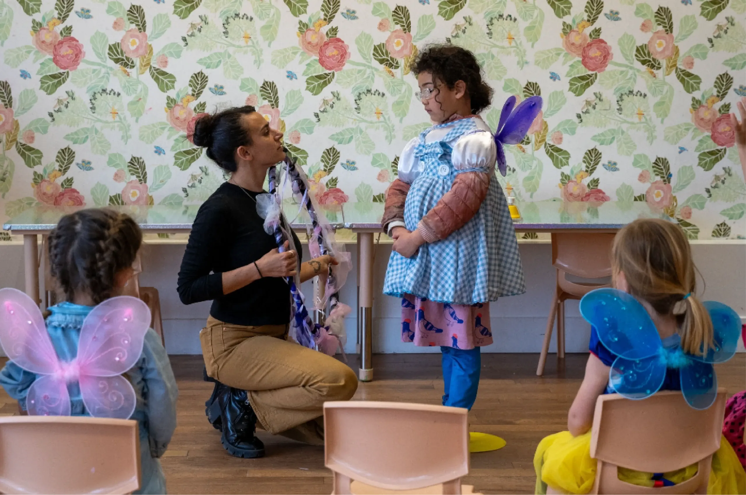 An adult kneels in front of a child dressed as a fairy, while two other children wearing wings sit nearby in a classroom with floral wallpaper.