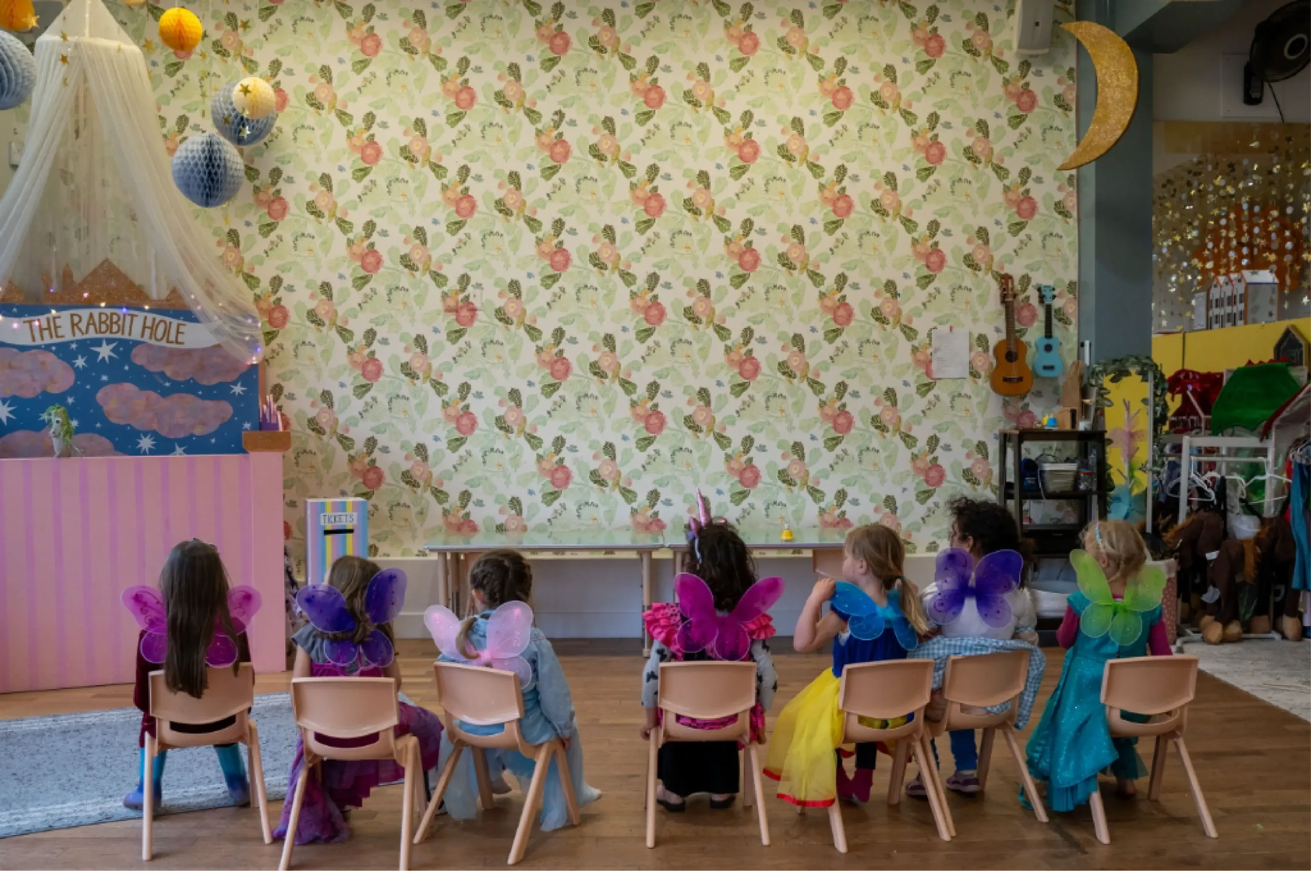 Seven children in costumes with colorful fairy wings sit on chairs facing a floral wallpapered wall in a decorated classroom.
