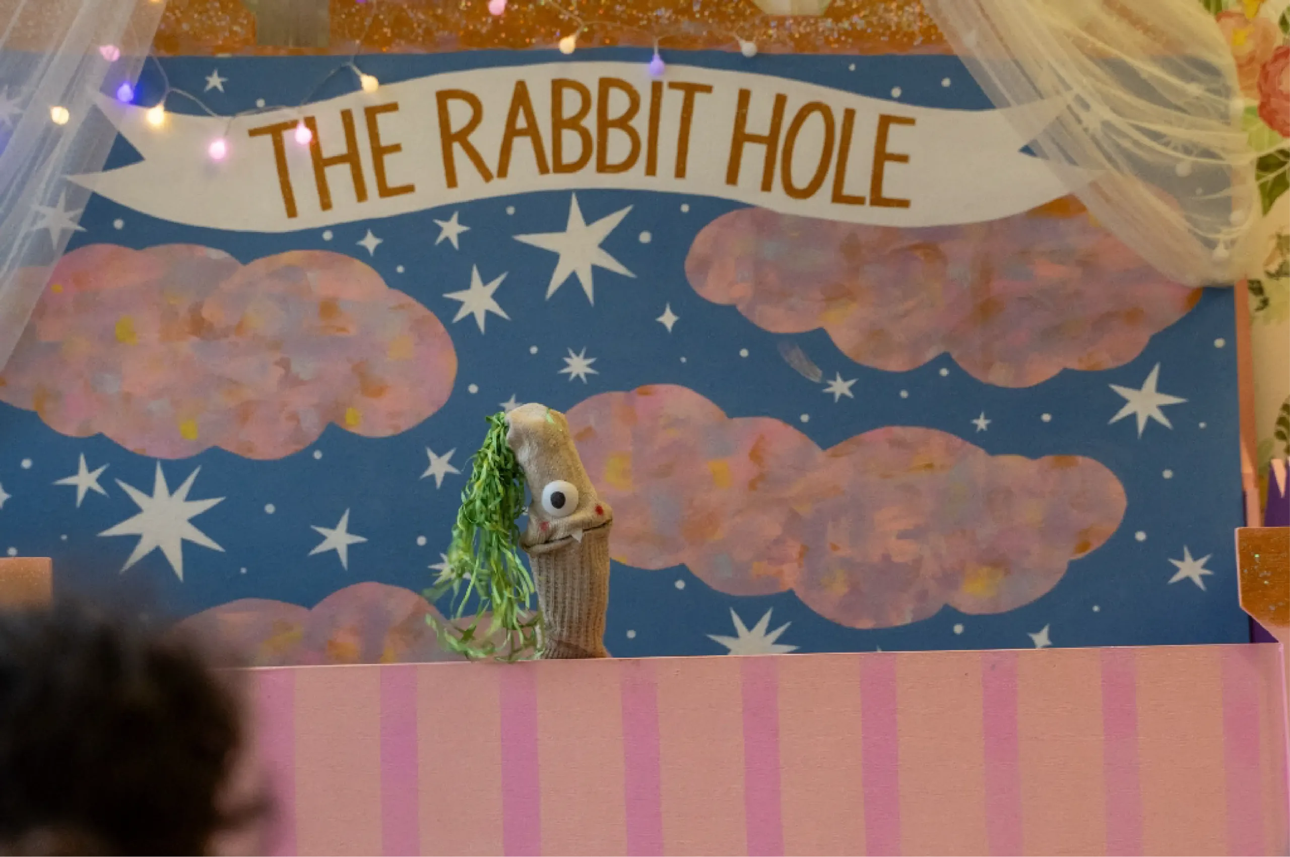 A puppet with googly eyes and green yarn hair is on stage under a sign that reads "The Rabbit Hole," with a painted backdrop of clouds and stars.